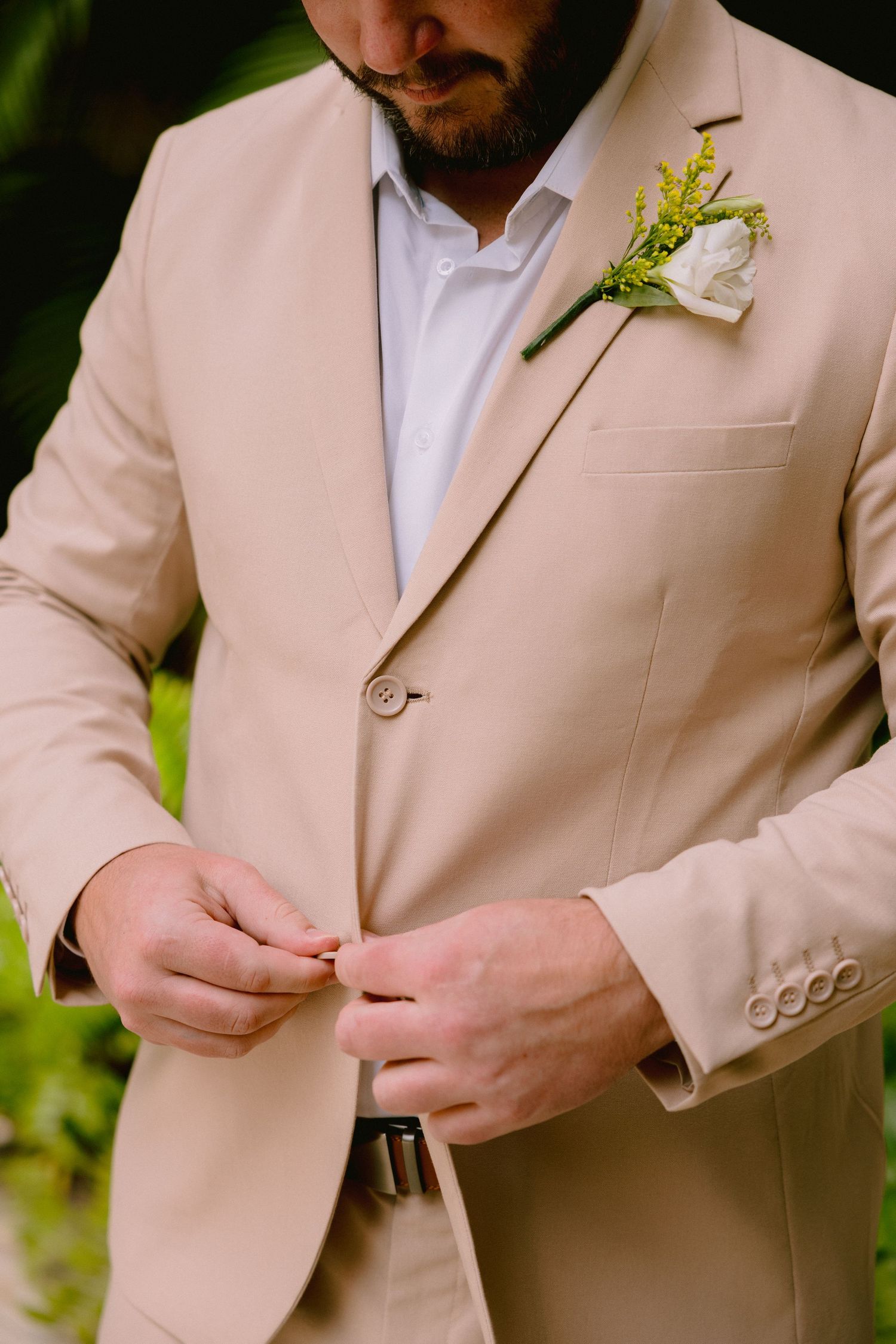 Groom in beige suit with white floral boutonniere buttoning jacket before wedding ceremony.