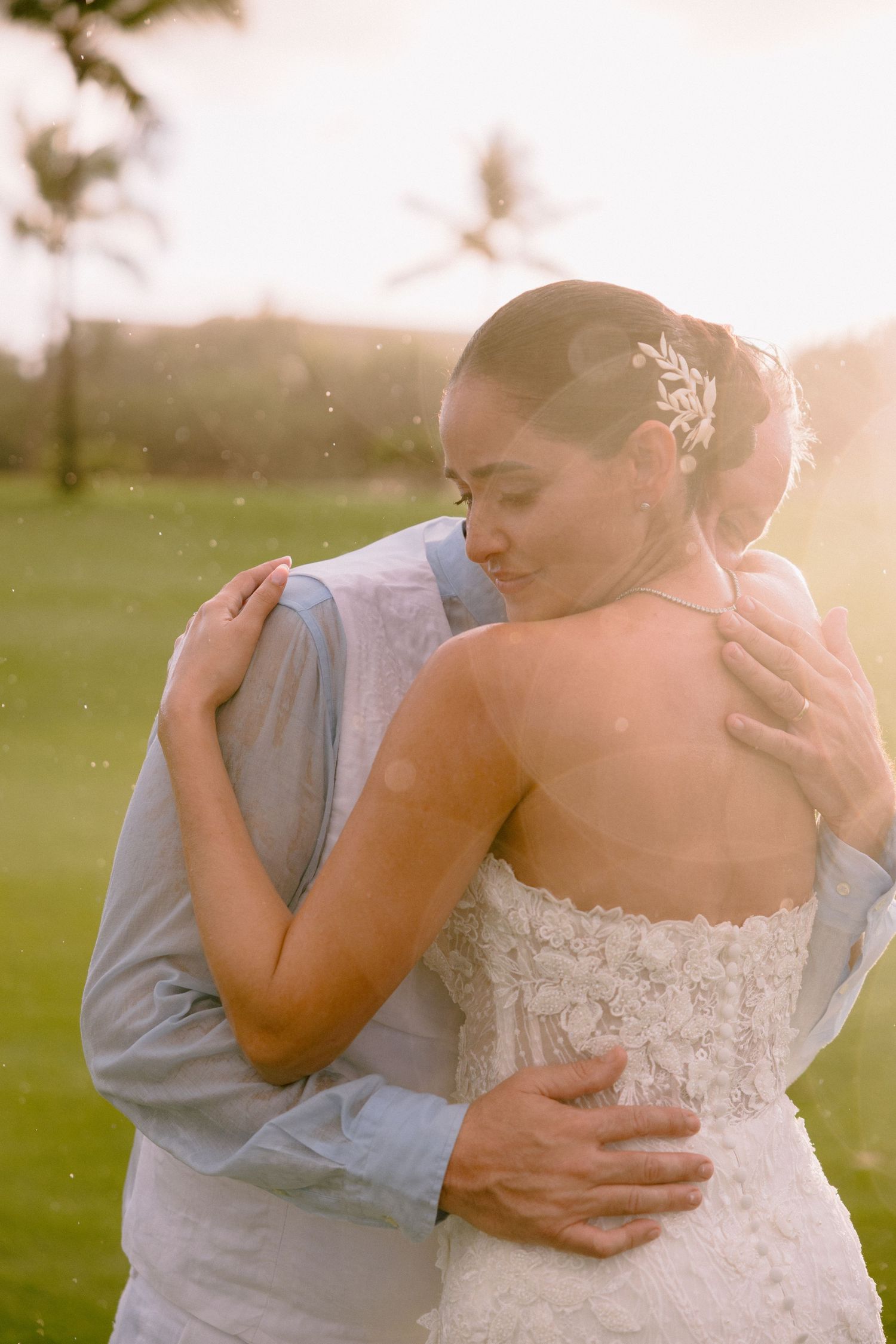 A couple in wedding attire share an intimate embrace during sunset on a grassy field.