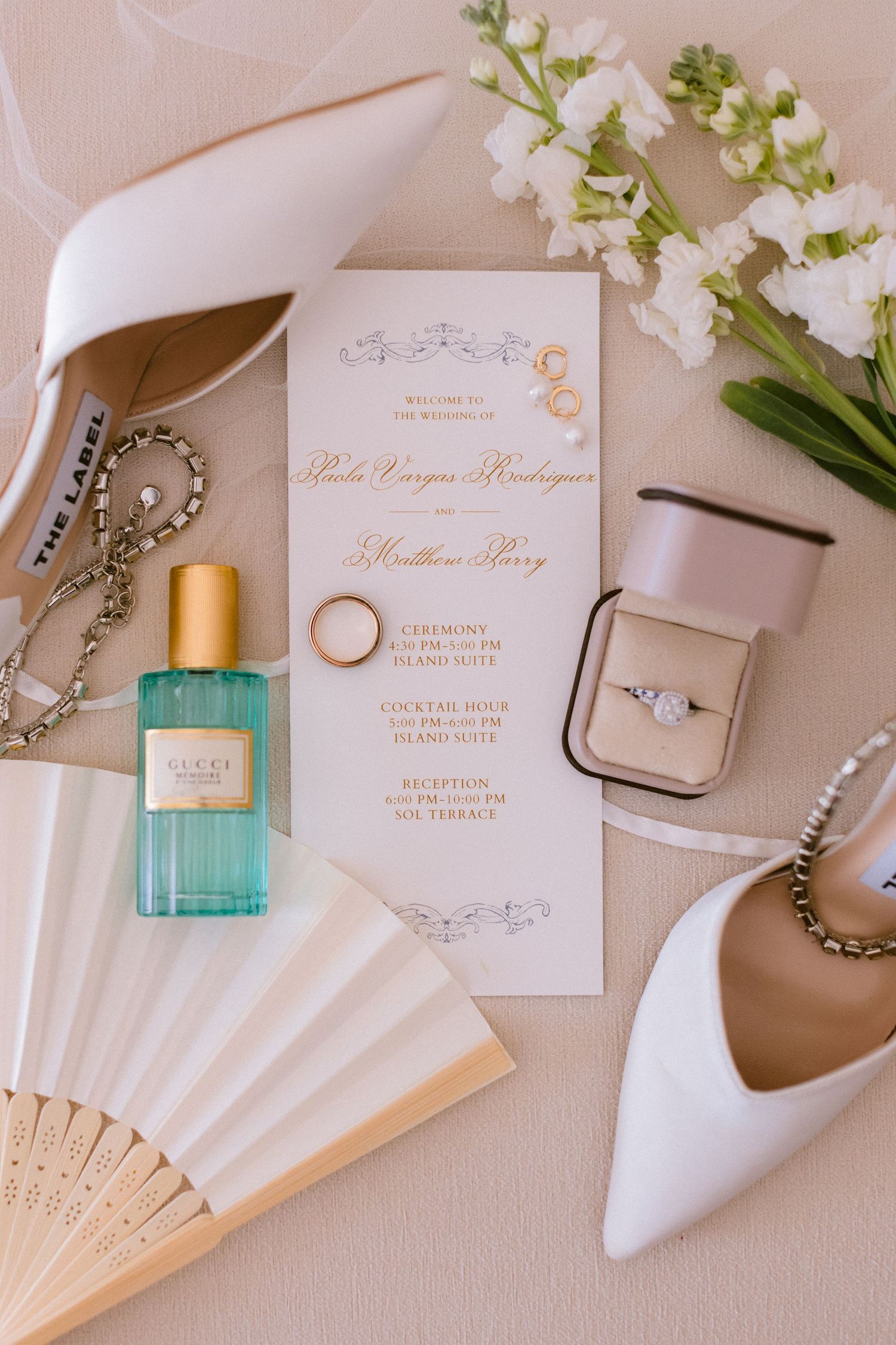 Elegant wedding details including white shoes, paper fan, turquoise perfume bottle, and gold calligraphy invitations on beige surface.