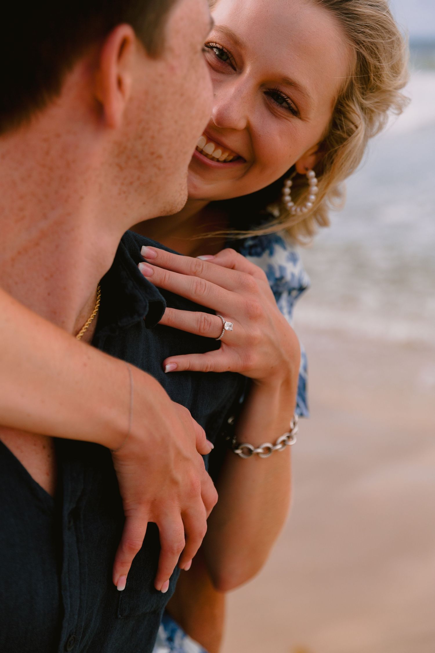 A close-up romantic moment between a couple on the beach, showing hands with an engagement ring.