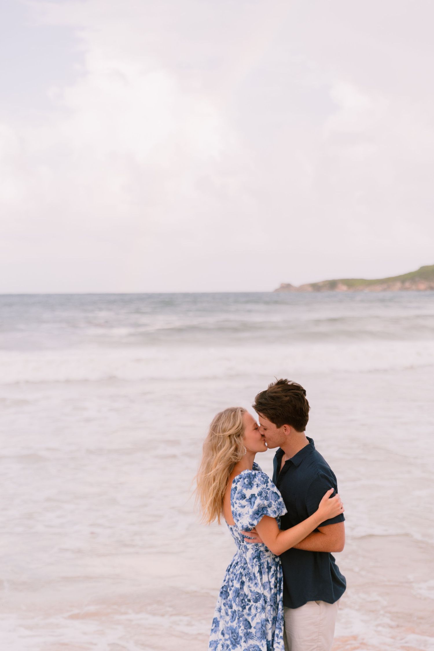 A romantic couple embraces on a misty beach with waves crashing in the background.