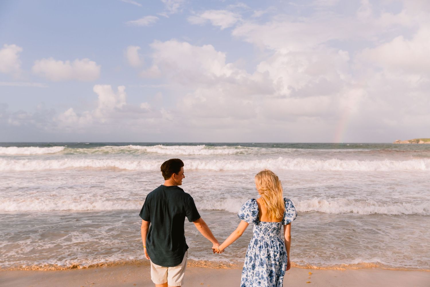 Two people walk hand in hand along a sandy beach with rolling waves and cloudy skies in the background.