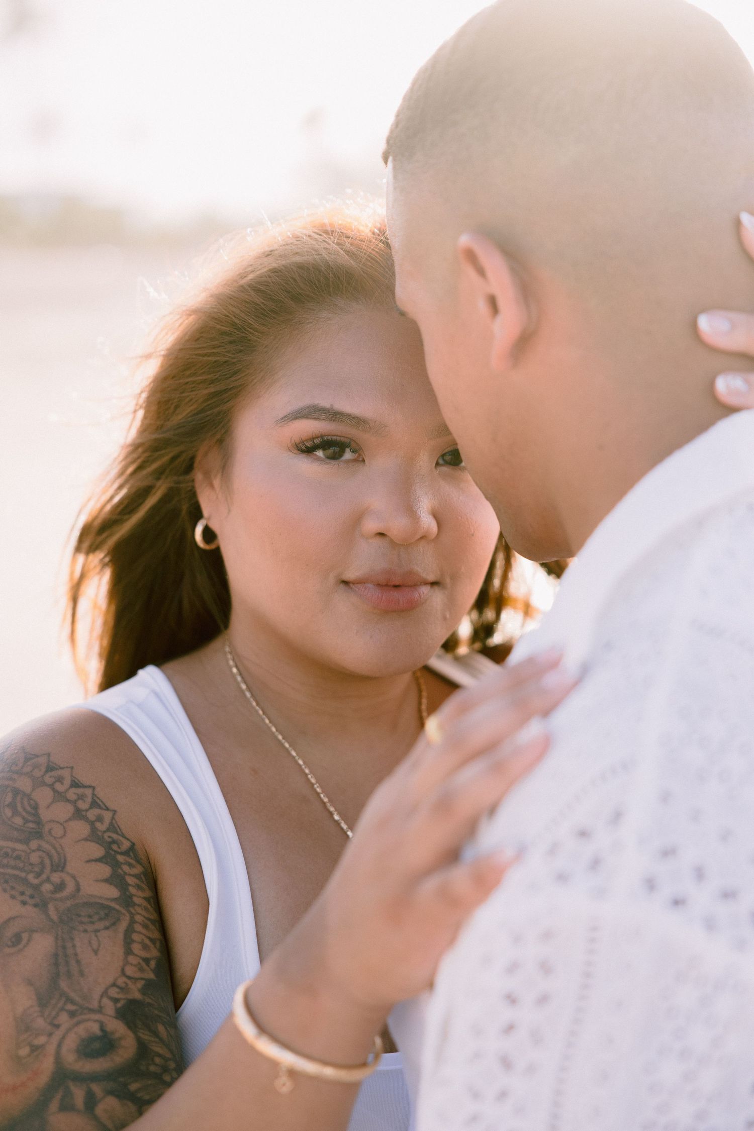 A woman with a tattoo and gold jewelry gazes at her partner during a golden hour beach engagement photo session.