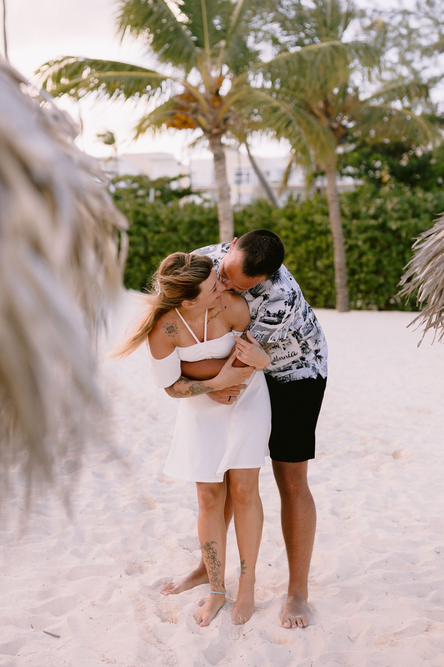 Couple sharing a romantic embrace on a white sand beach surrounded by palm trees.