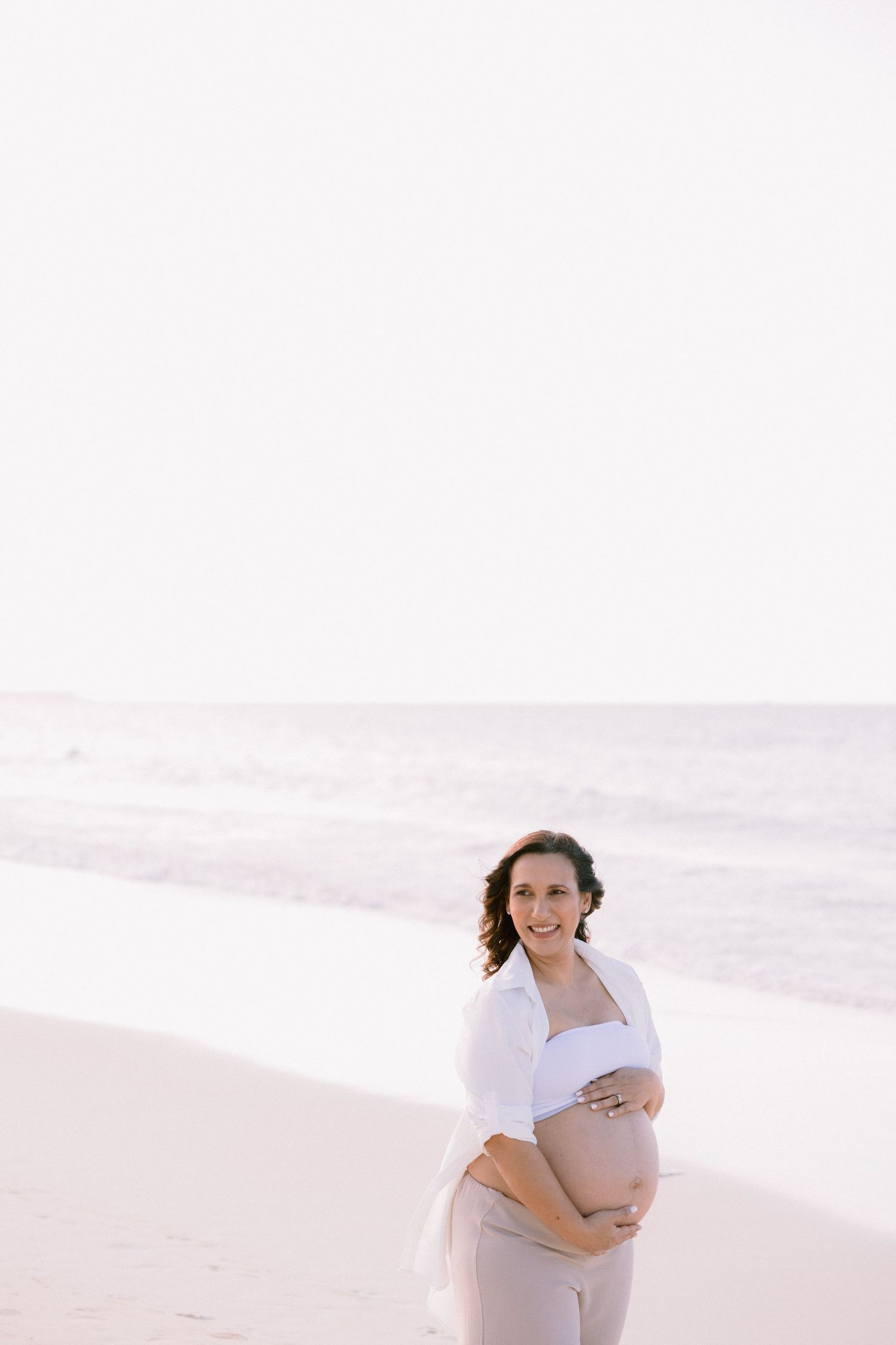 Smiling pregnant woman in white top holds baby bump on a bright sandy beach.