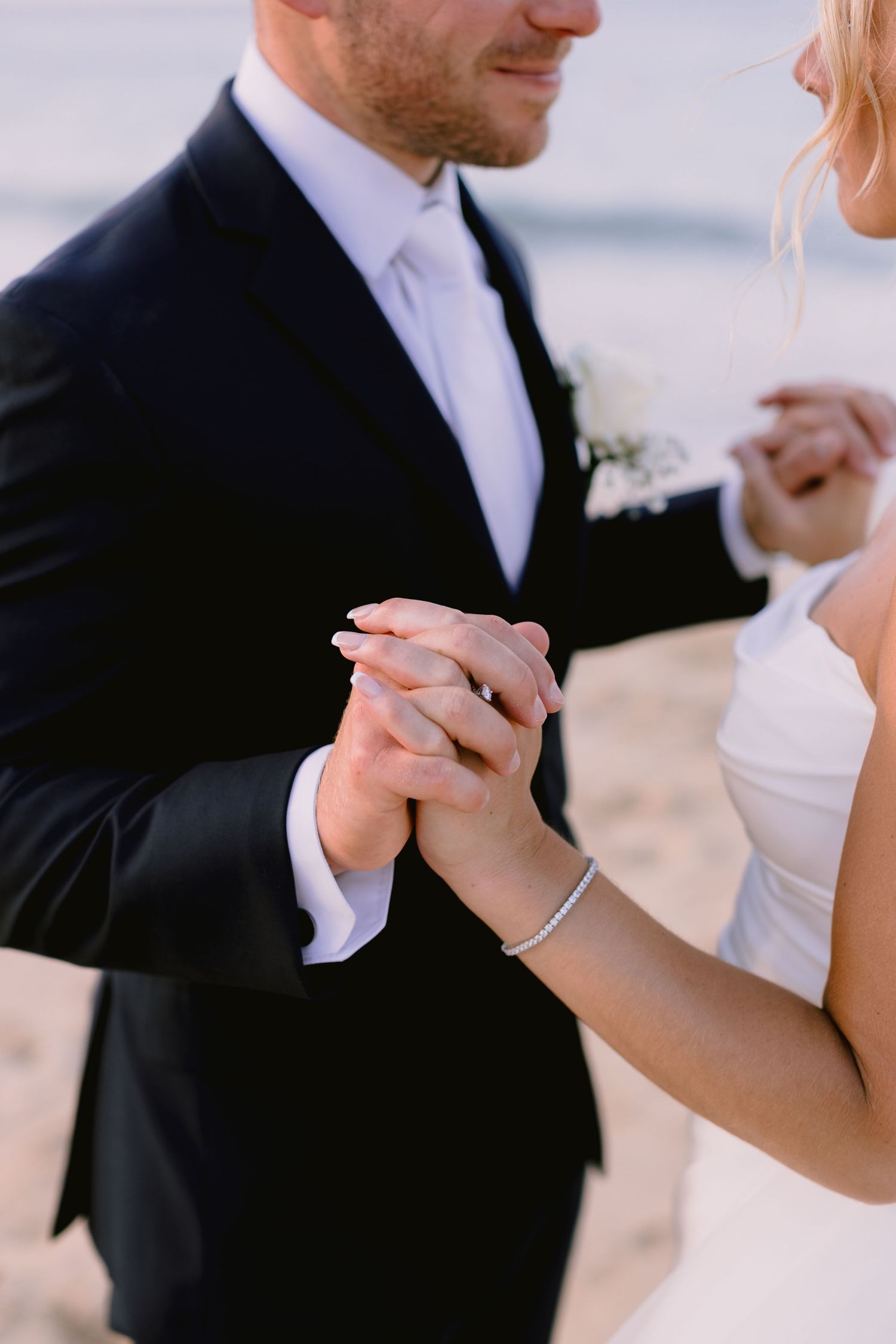 Bride and groom holding hands at beach wedding, bride wearing diamond bracelet, groom in dark suit with white boutonniere.