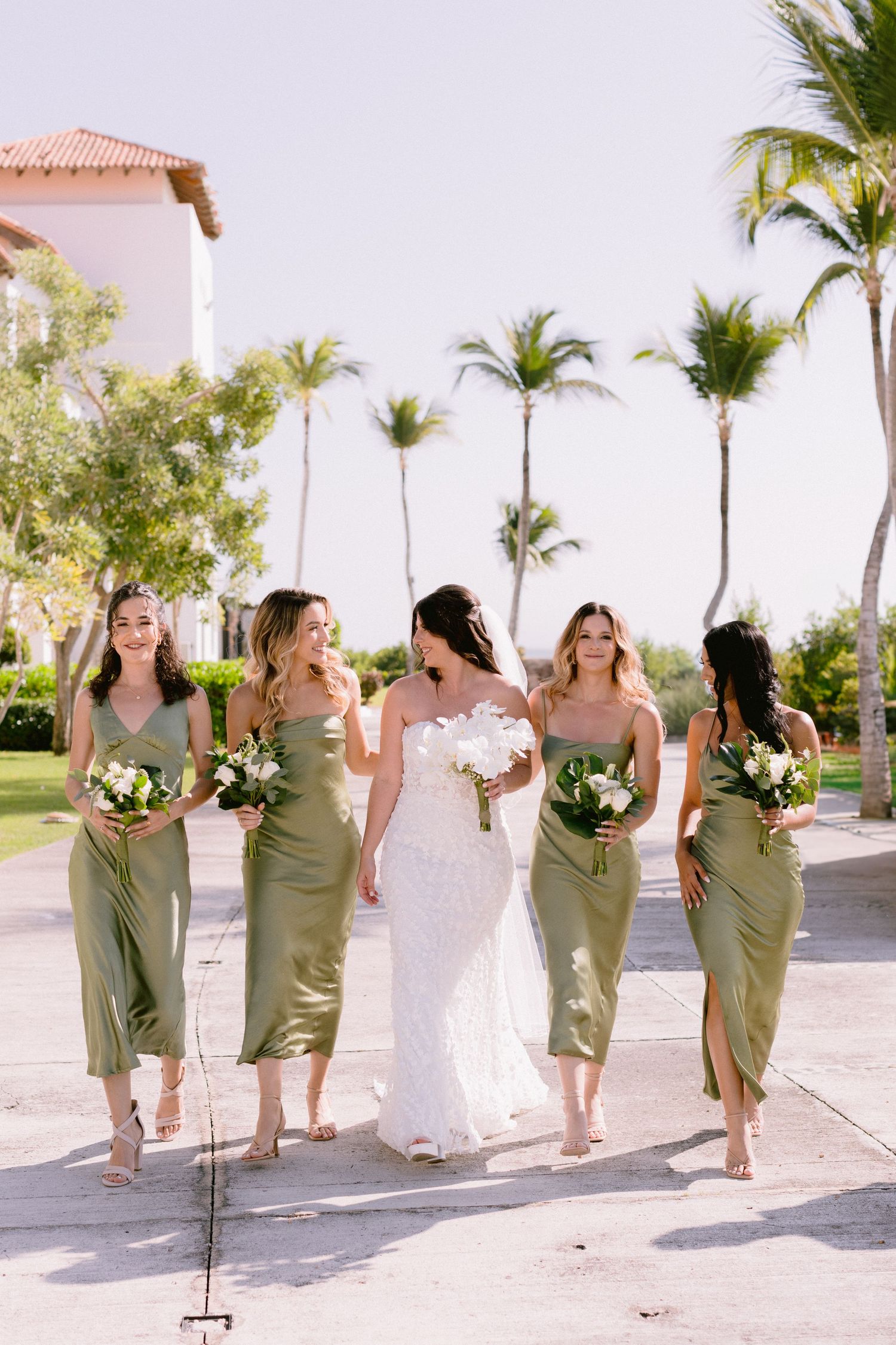 A wedding party in sage green dresses and white gown walks along a tropical palm-lined path during a destination ceremony.