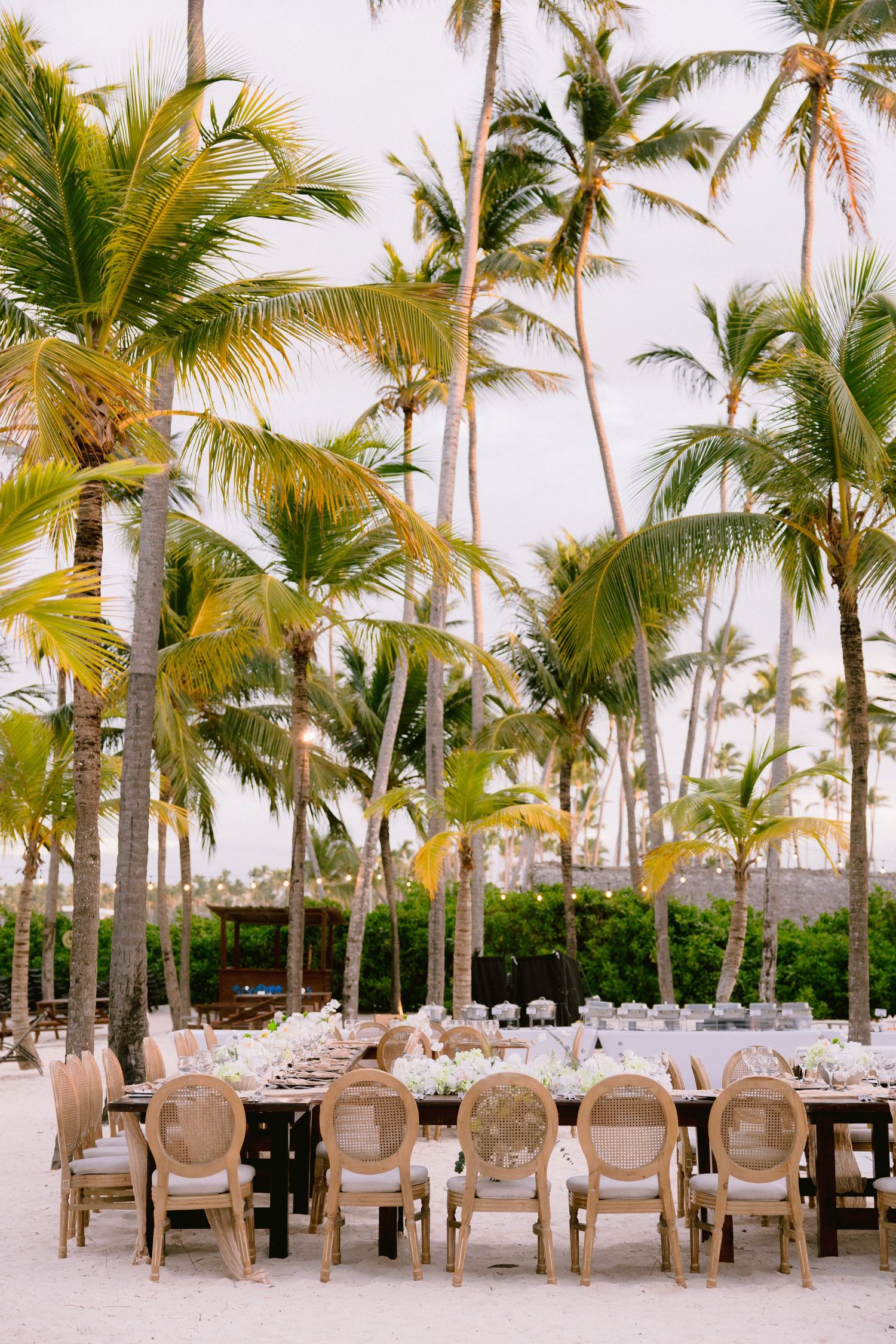 Elegant outdoor beach wedding reception with rattan chairs and long dining tables set among tall tropical palm trees.