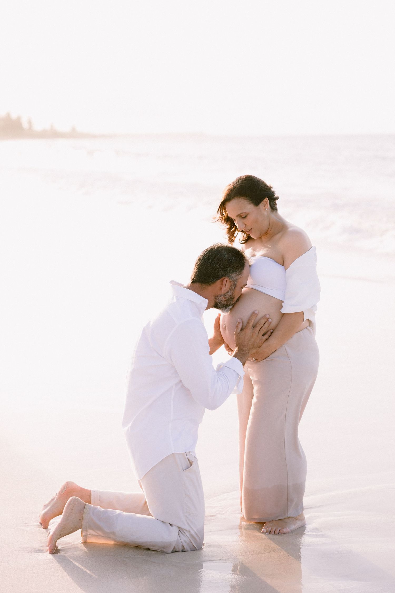 Man kneeling on beach kissing pregnant woman's belly during golden hour maternity photoshoot.