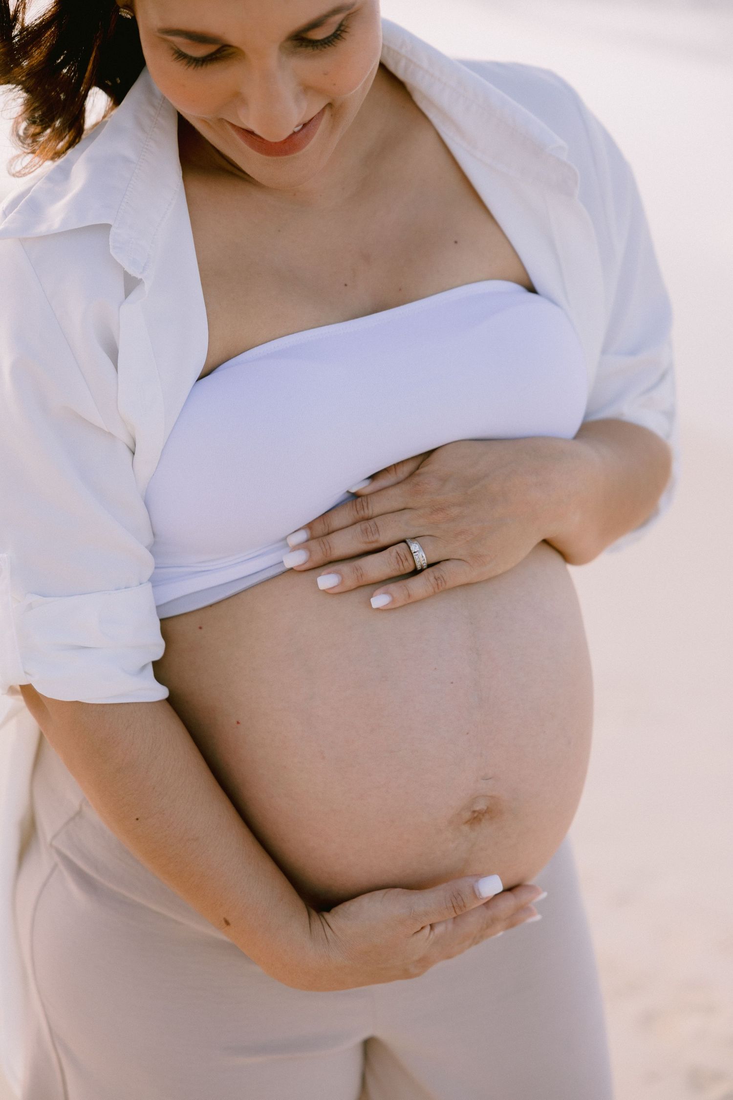 Pregnant woman in white crop top and open shirt cradling her baby bump on a sunny beach.