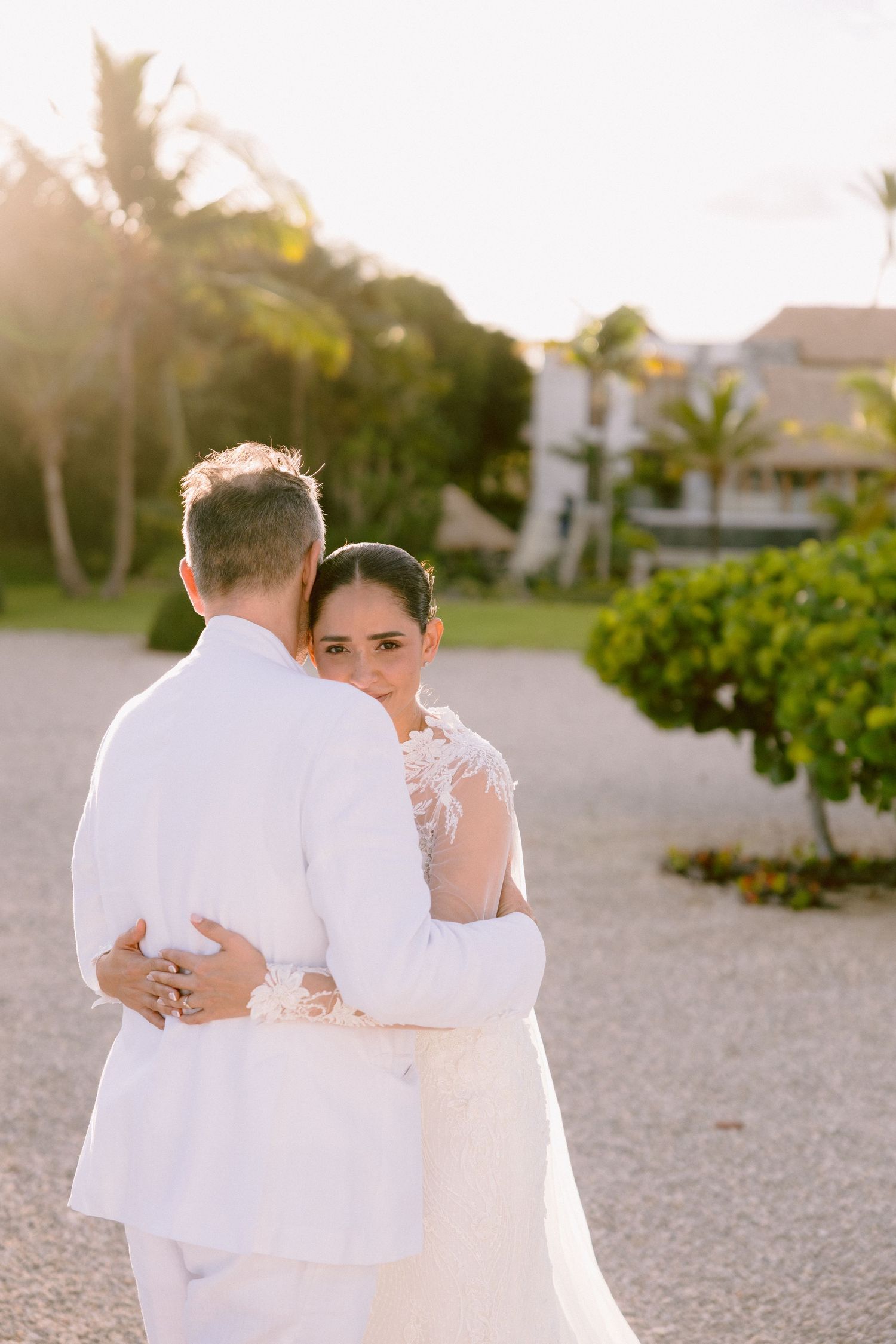 A couple in white attire embraces on a sandy beach path during sunset with lush greenery in the background.