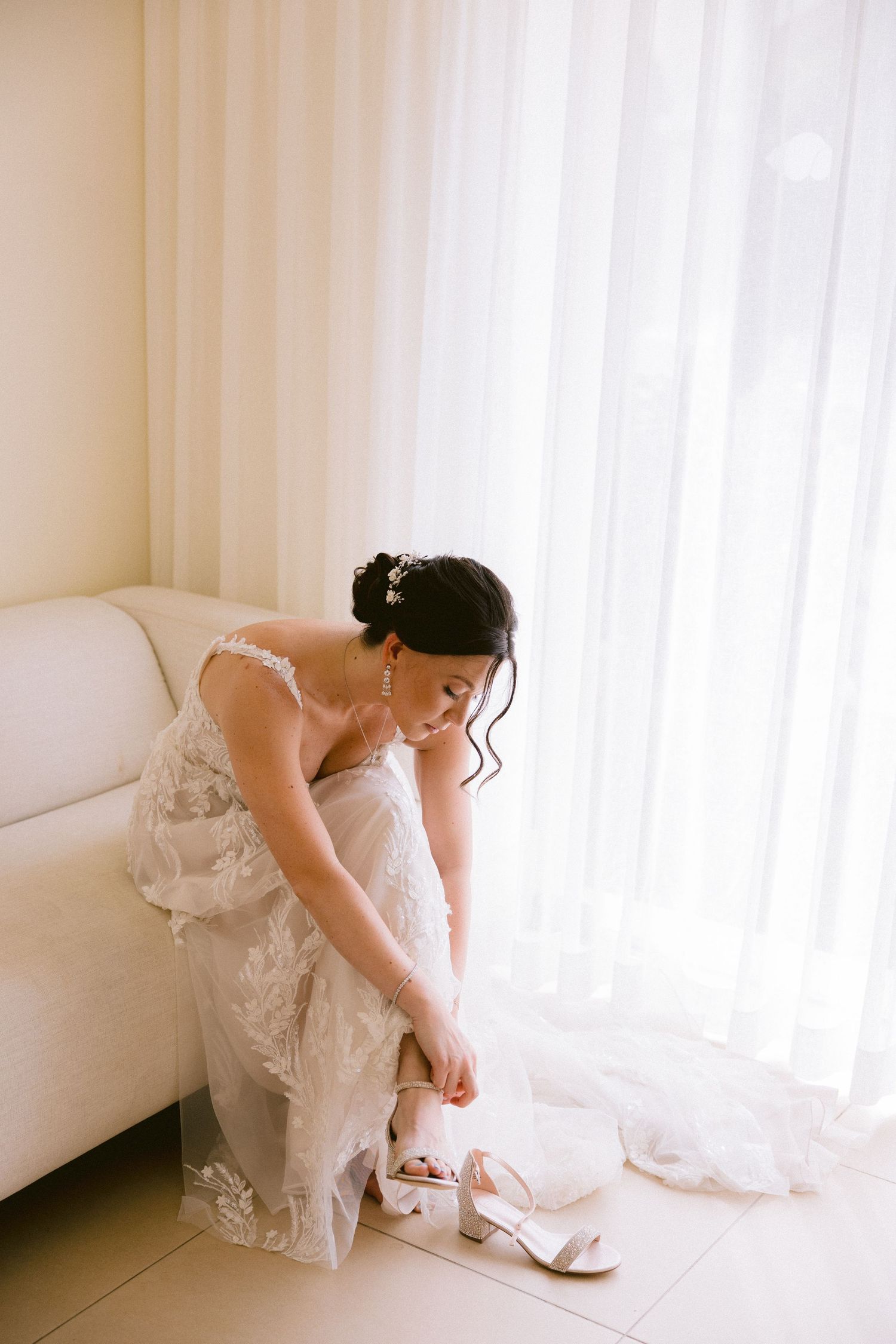 A bride in a white wedding dress adjusts her elegant heels by a bright window in a soft, dreamy setting.