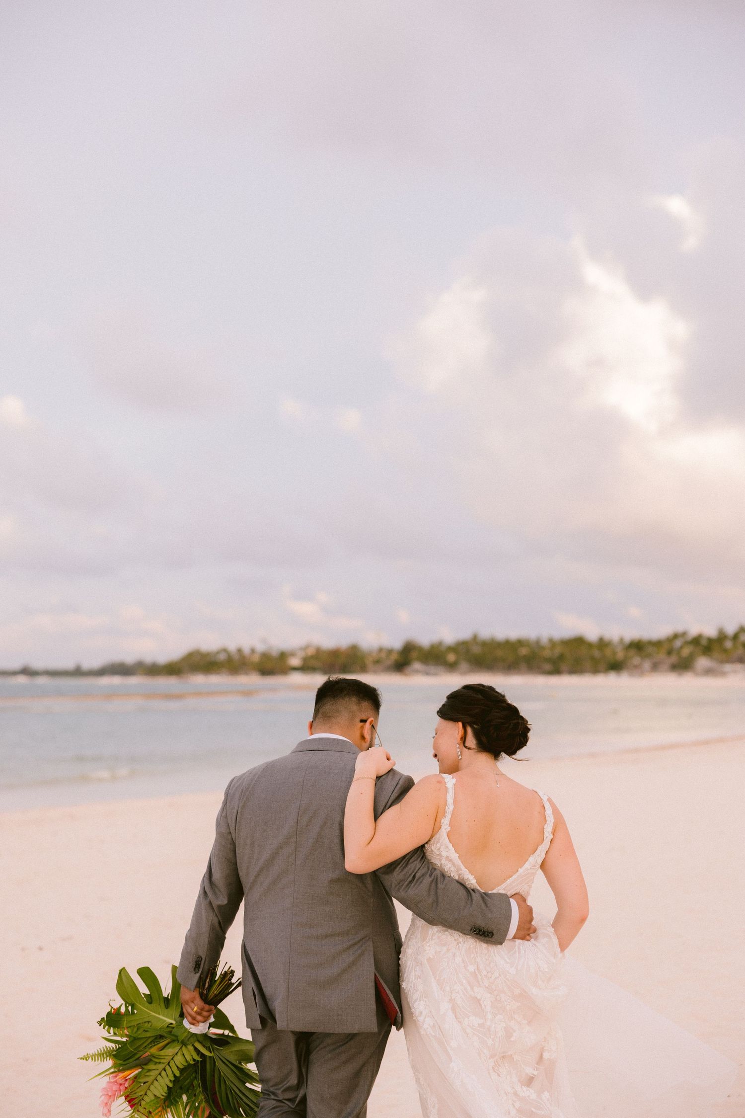 A couple walks along a tropical beach at sunset with palm fronds in the foreground.