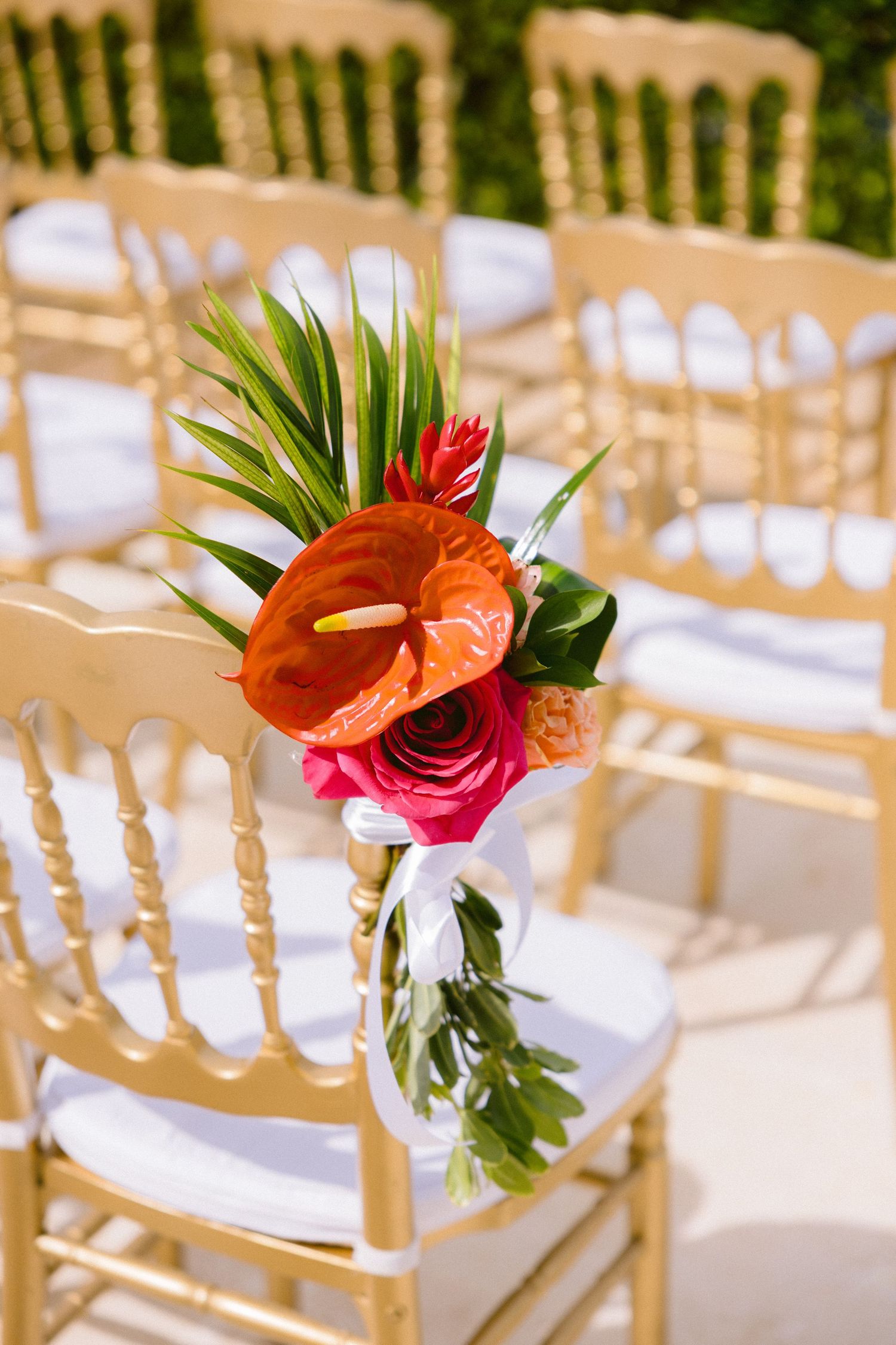 Tropical flowers with anthurium and roses decorate white chairs at an outdoor wedding ceremony setup.