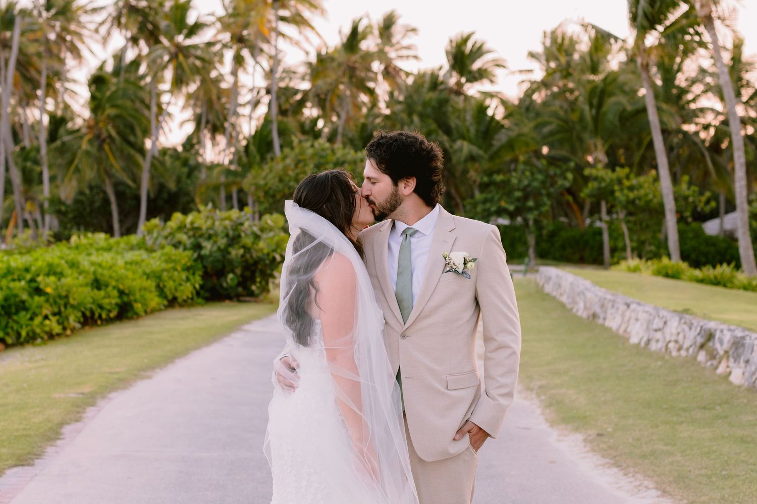 Couple in elegant wedding attire embraces while walking along a palm-lined tropical beach path at sunset.