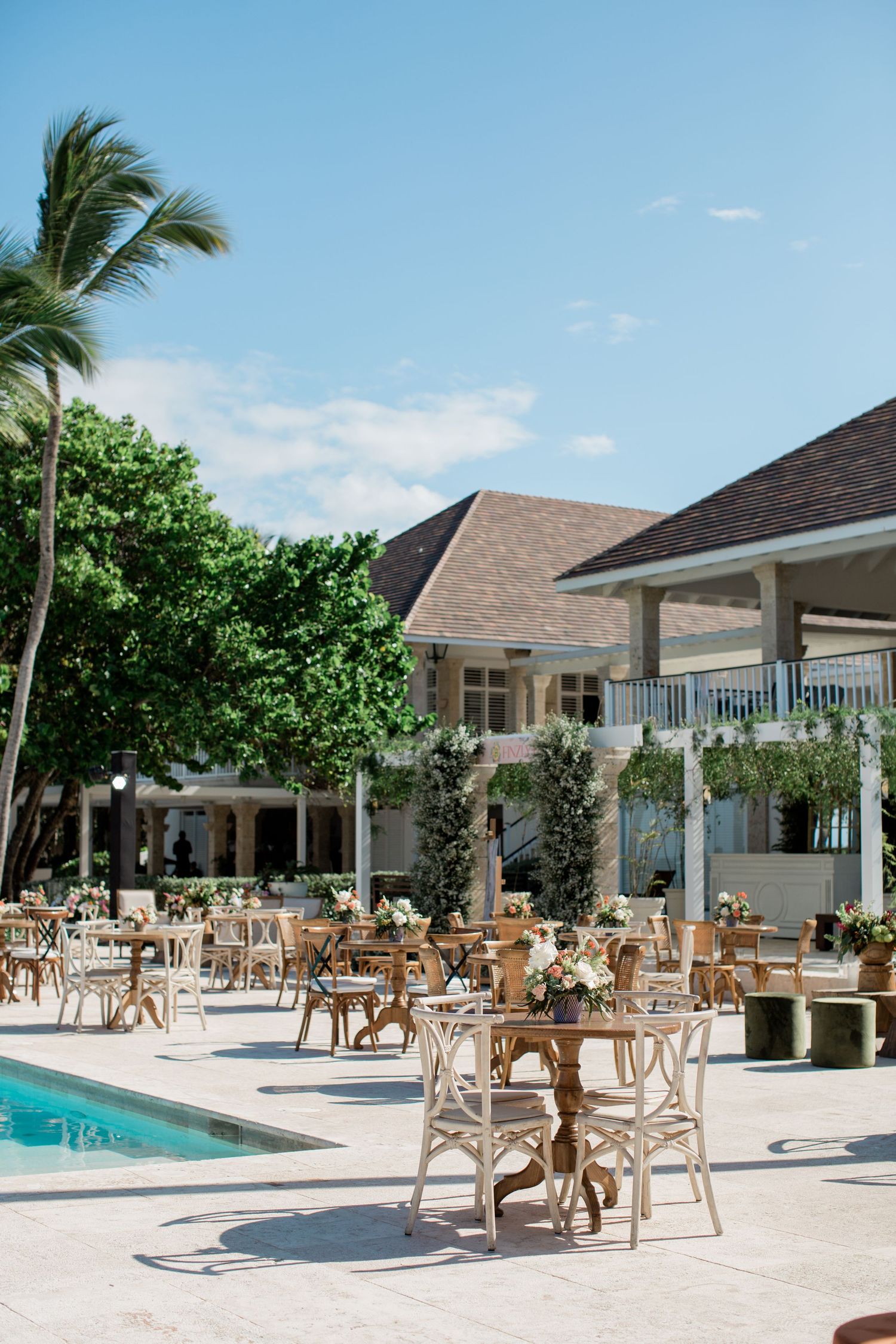 Tropical resort poolside dining area with rattan furniture, palm trees and white colonial-style building under blue sky.