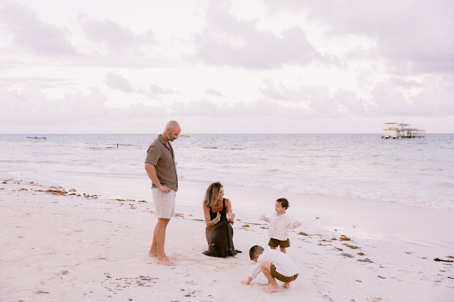 Family of four enjoying a relaxing day on a white sand beach with calm ocean waters and a boat in the background.