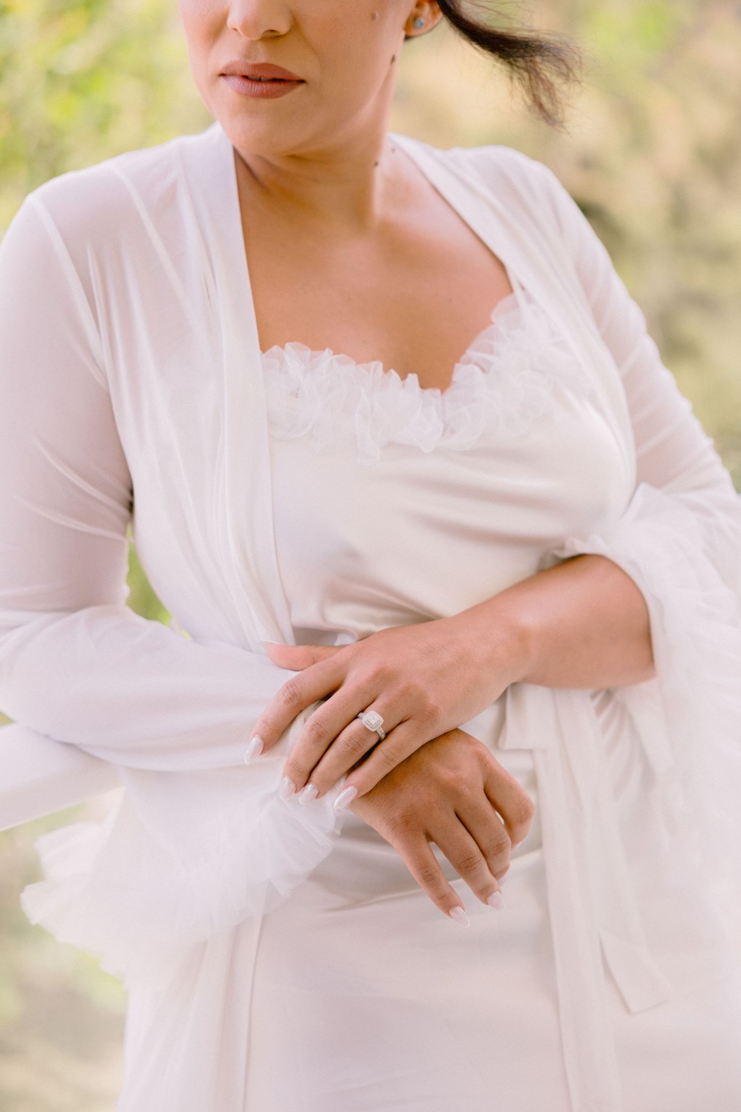 Bride in white robe adjusting sleeve, showcasing elegant diamond engagement ring, with soft natural background.