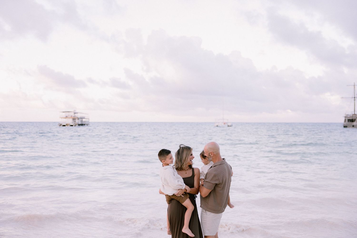 Couple enjoys a romantic moment on a misty beach with boats visible in the distance at sunset.
