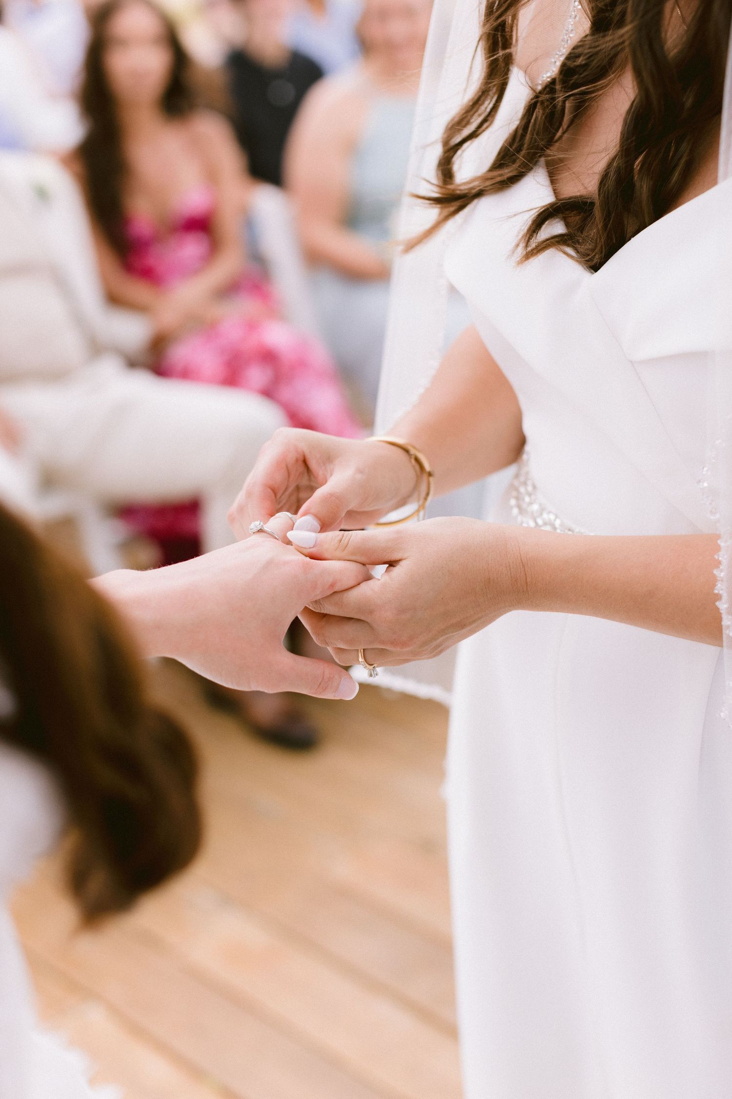 Bride and groom exchanging wedding rings during ceremony, guests seated in background, bride wearing white dress and gold bracelet.