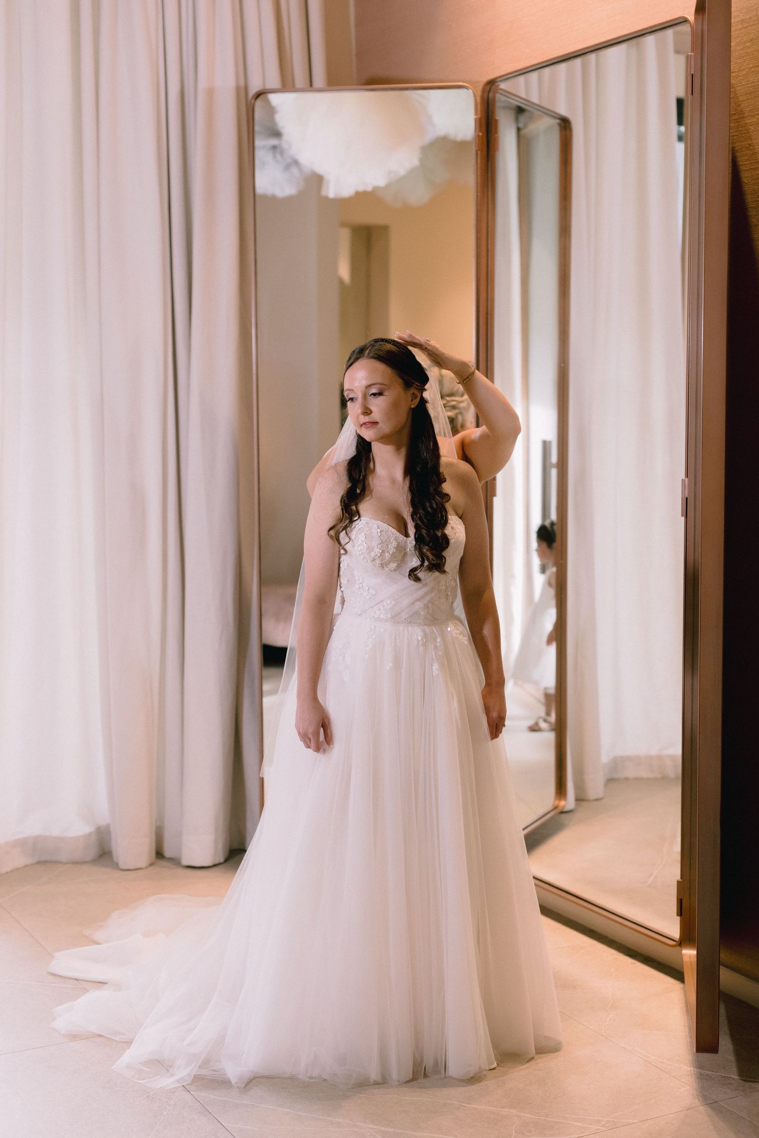 Bride in strapless white tulle wedding gown poses in bridal boutique fitting room with mirrors.
