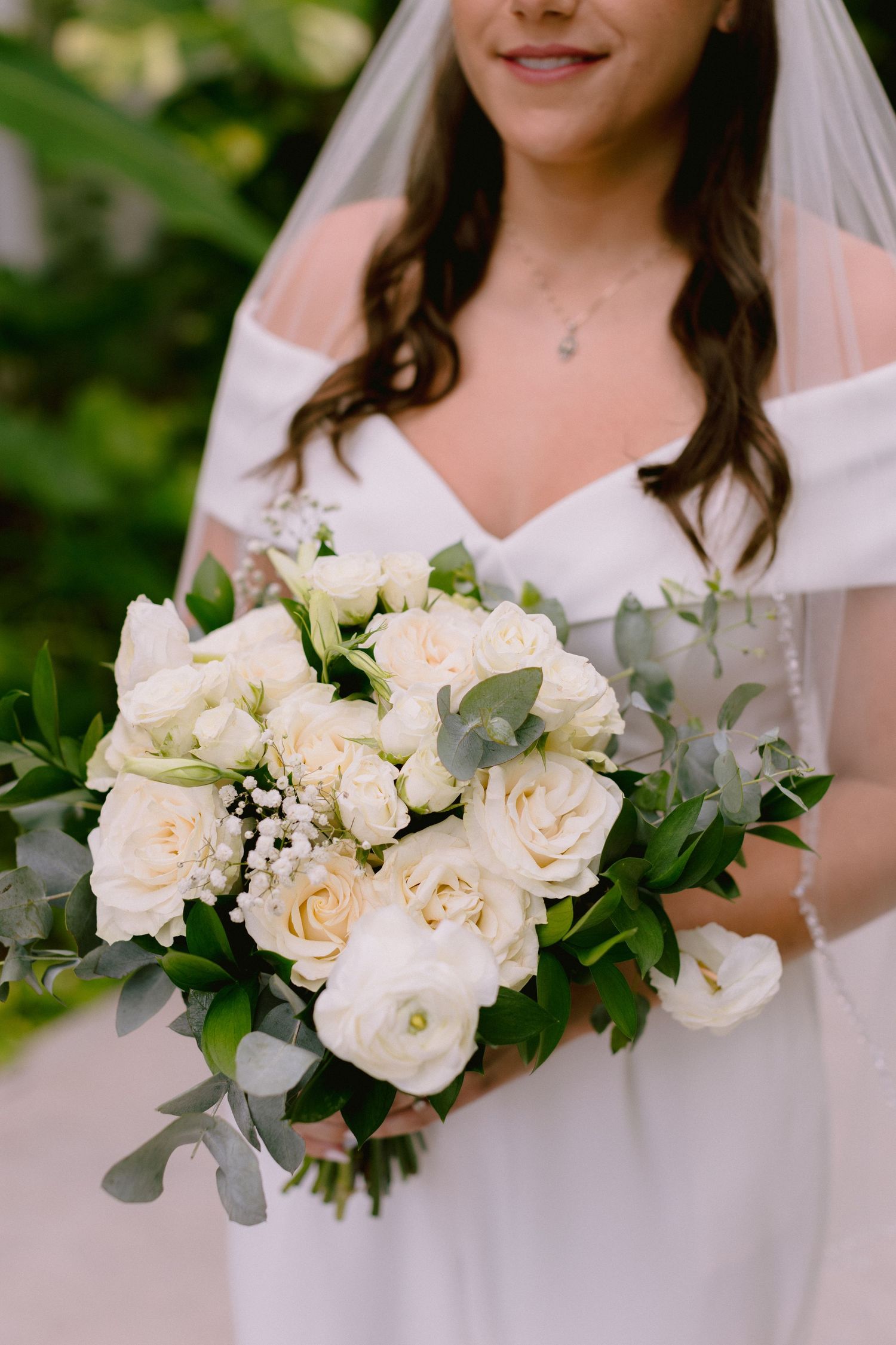 Bride in white off-shoulder gown holding elegant bouquet of white roses and greenery on her wedding day.
