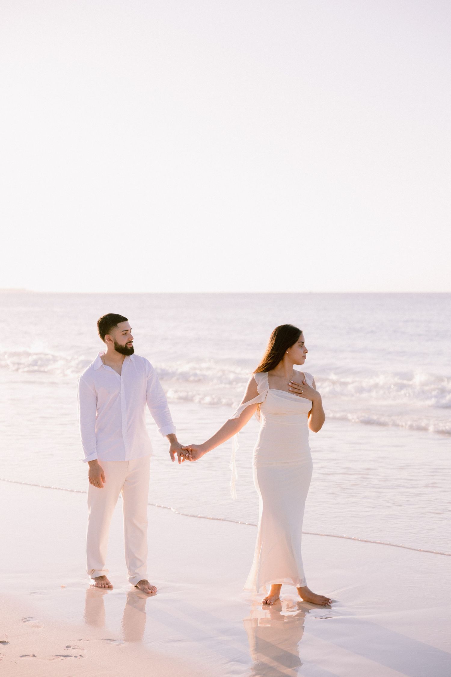 A couple in white outfits holds hands while walking barefoot on a serene beach at sunset.