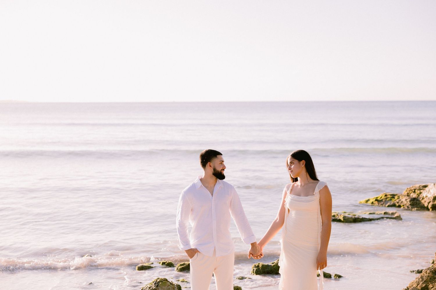 Couple in white outfits holding hands on a rocky beach with ocean waves in the background.