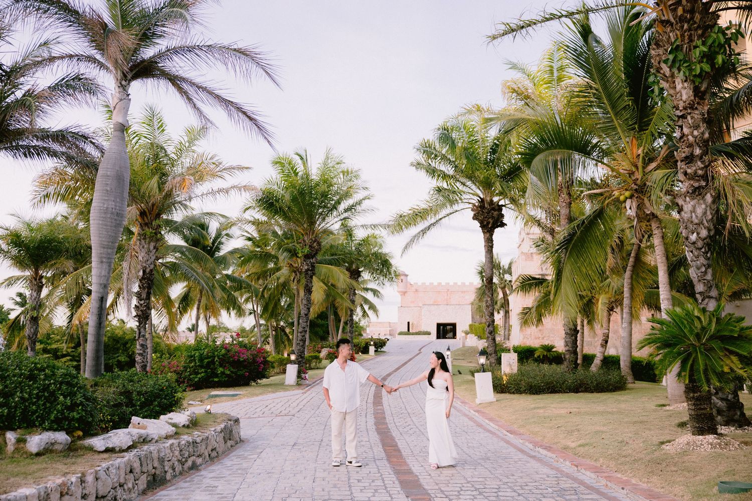 Couple holding hands on a tropical resort pathway surrounded by lush palm trees at sunset.