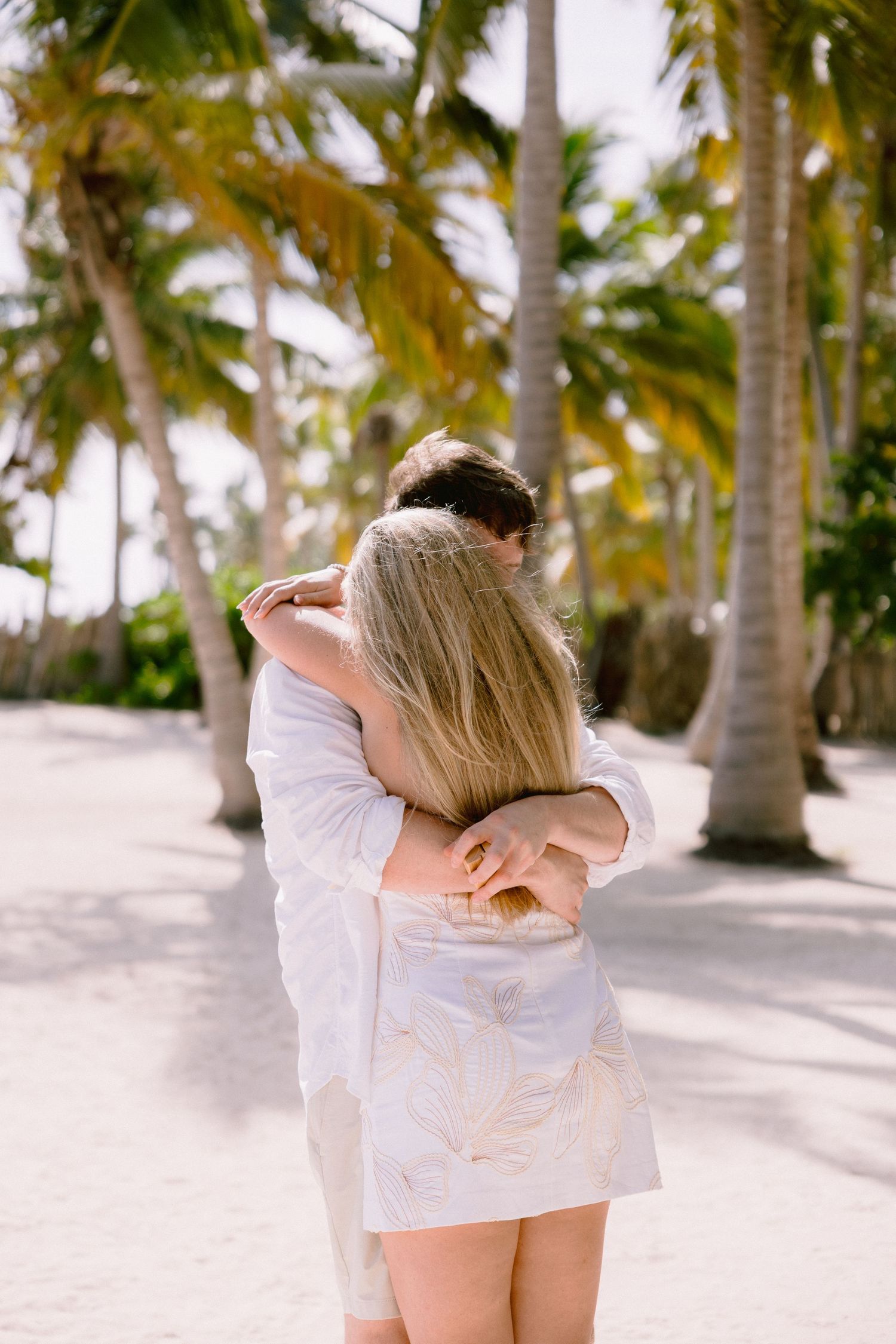 A couple embraces on a tropical beach with tall palm trees swaying in the background on a sunny day.
