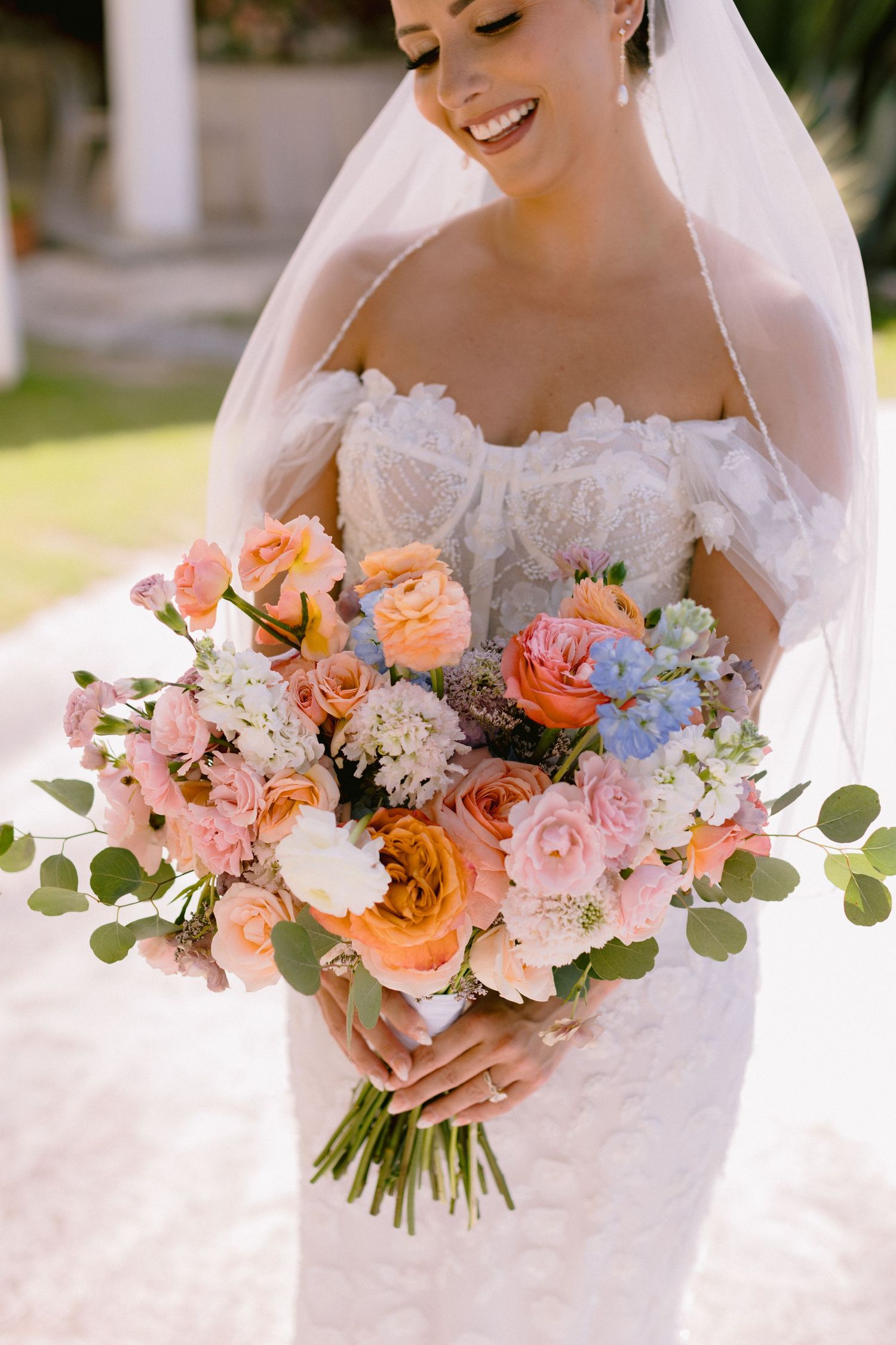 Smiling bride in lace off-shoulder gown holding a lush pastel bouquet of pink, orange, and blue flowers with eucalyptus.