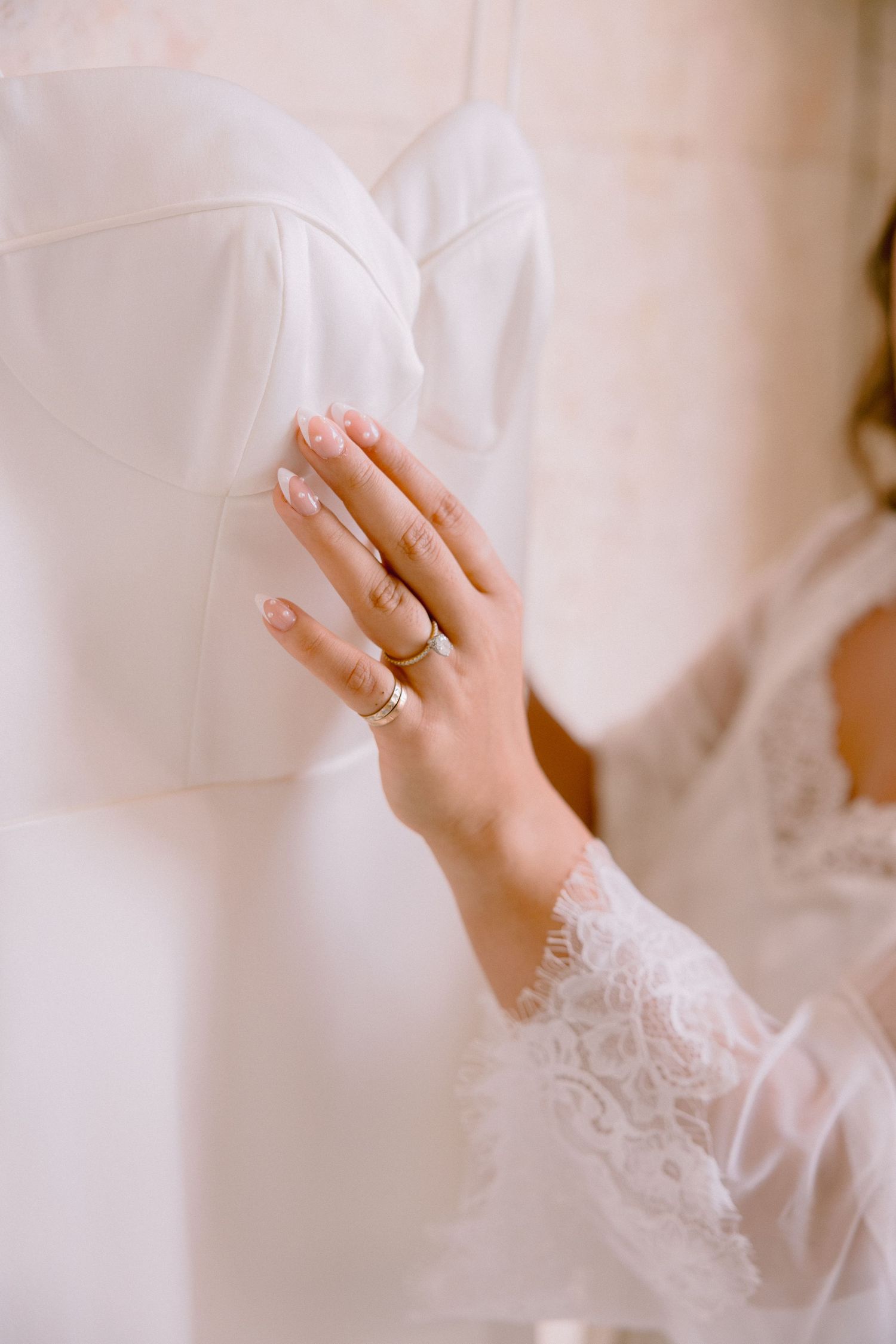 Bride's hand with gold rings touching white wedding dress, wearing lace robe sleeve, elegant bridal preparation moment.