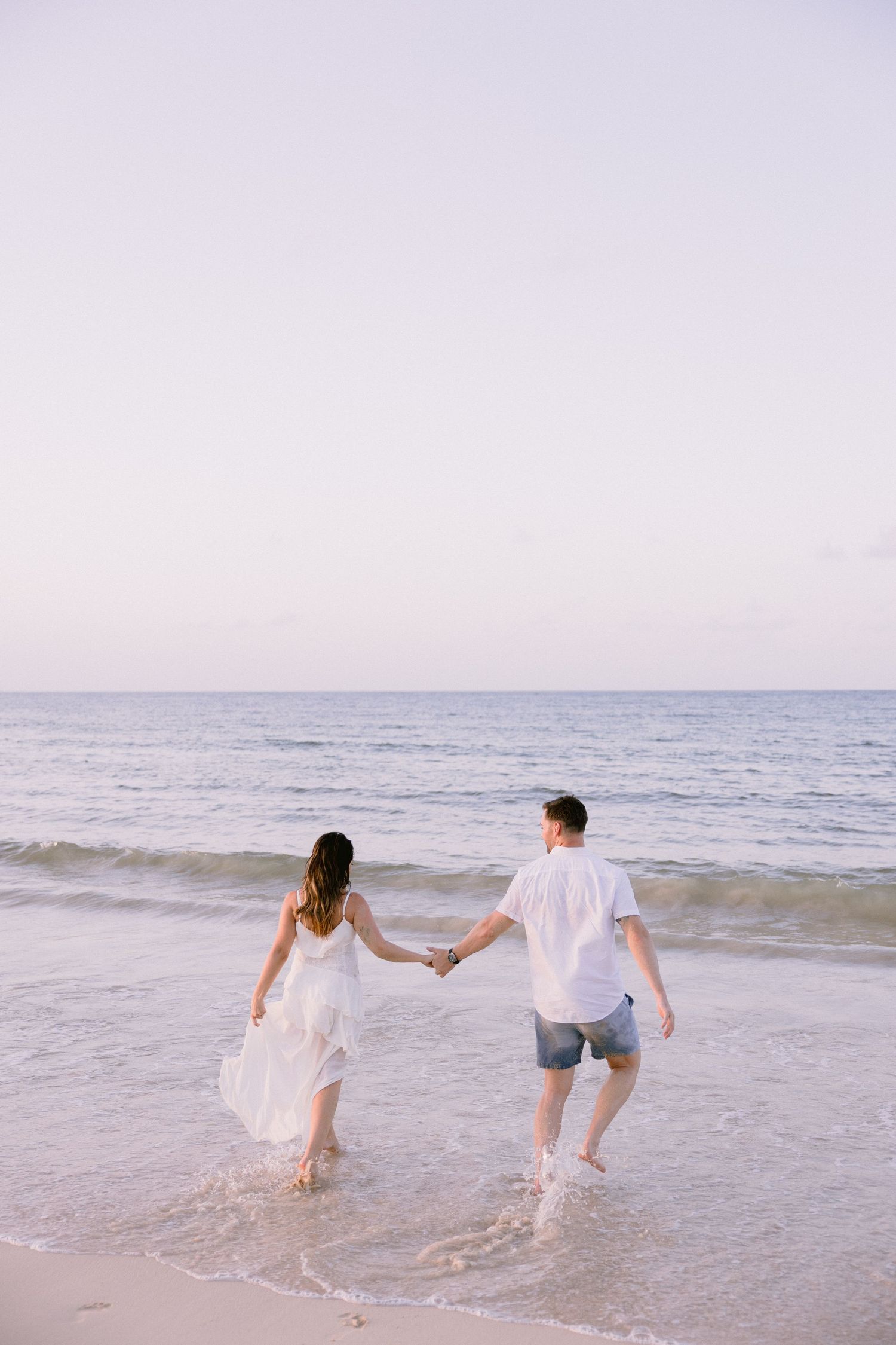 Couple holding hands walking into ocean waves at sunset, woman in white dress and man in white shirt and blue shorts.
