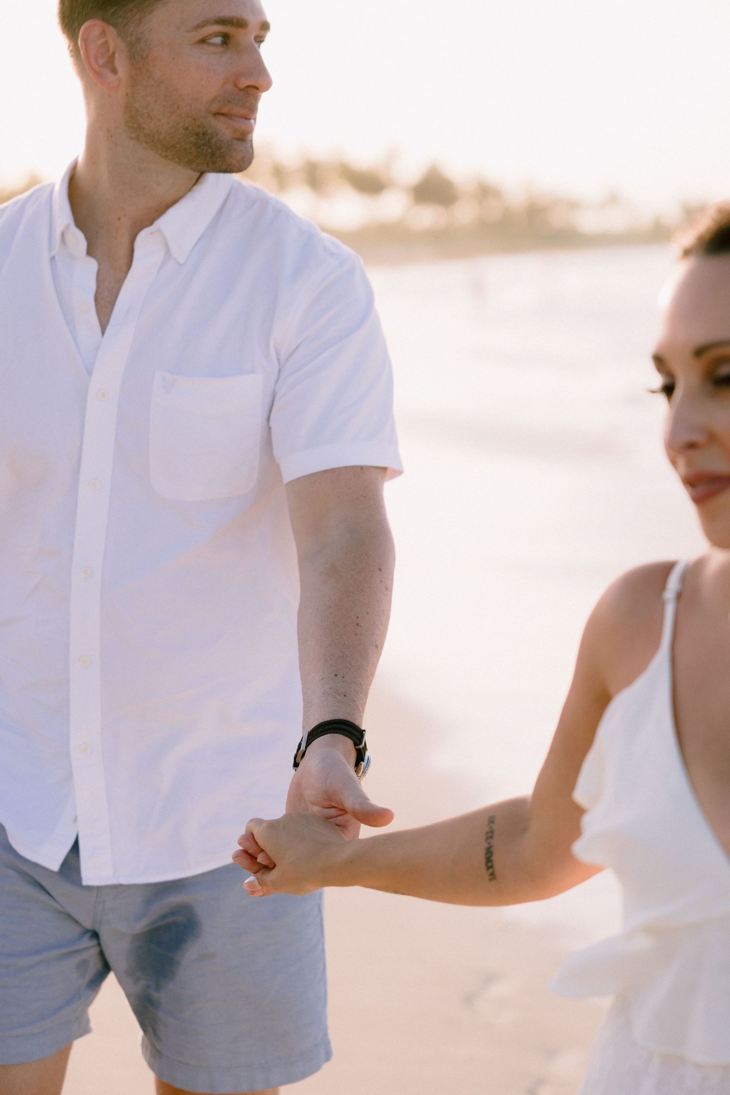 Couple holding hands on a sunny beach, man in white shirt and blue shorts, woman in white dress.