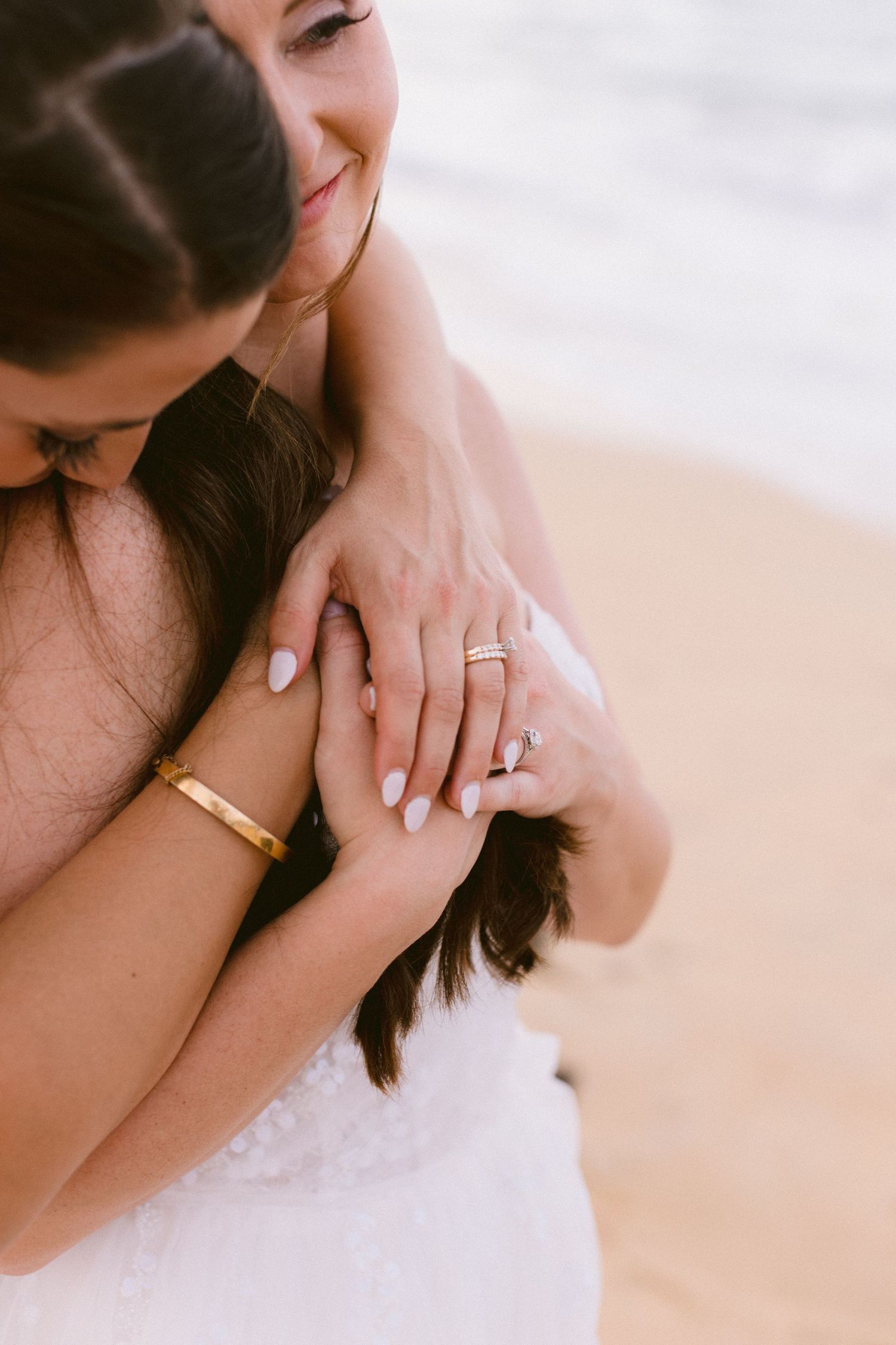 Two brides embracing on beach, showing wedding rings and gold bracelet, wearing white lace dress.