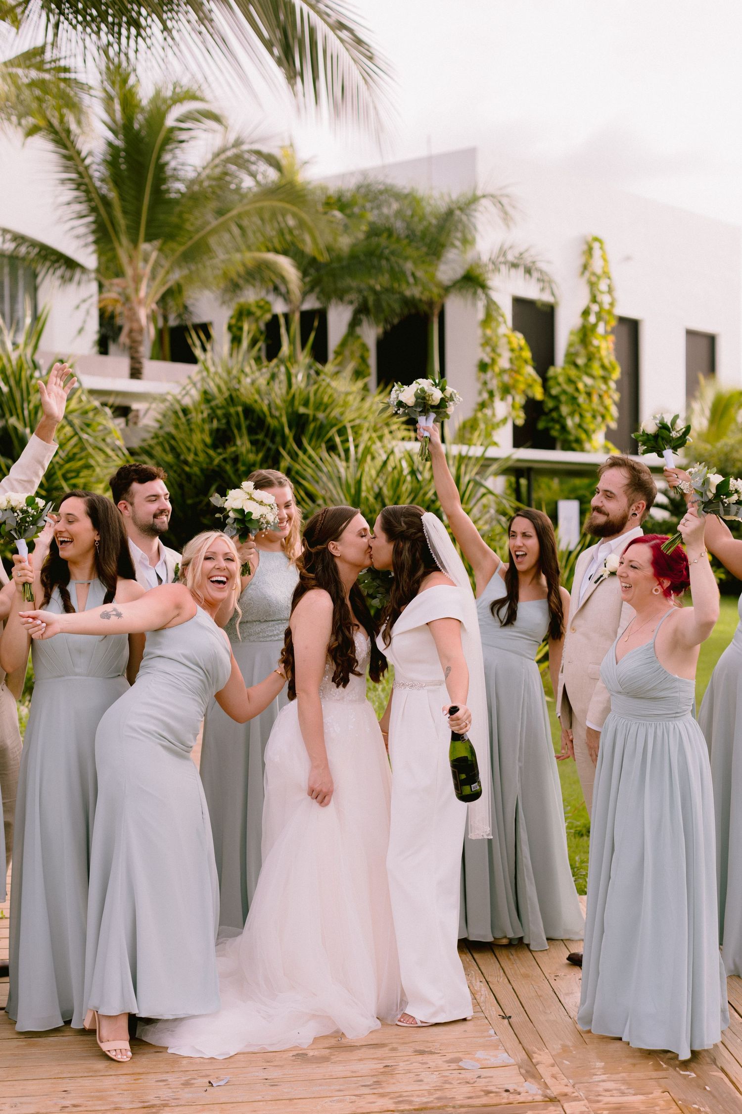Two brides share a kiss surrounded by bridesmaids in sage green gowns at a tropical outdoor wedding celebration.