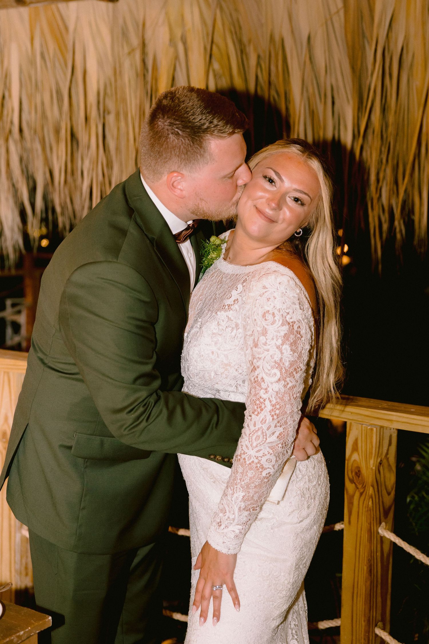 Groom in green suit kisses bride in lace dress on the cheek as she smiles at their wedding reception.