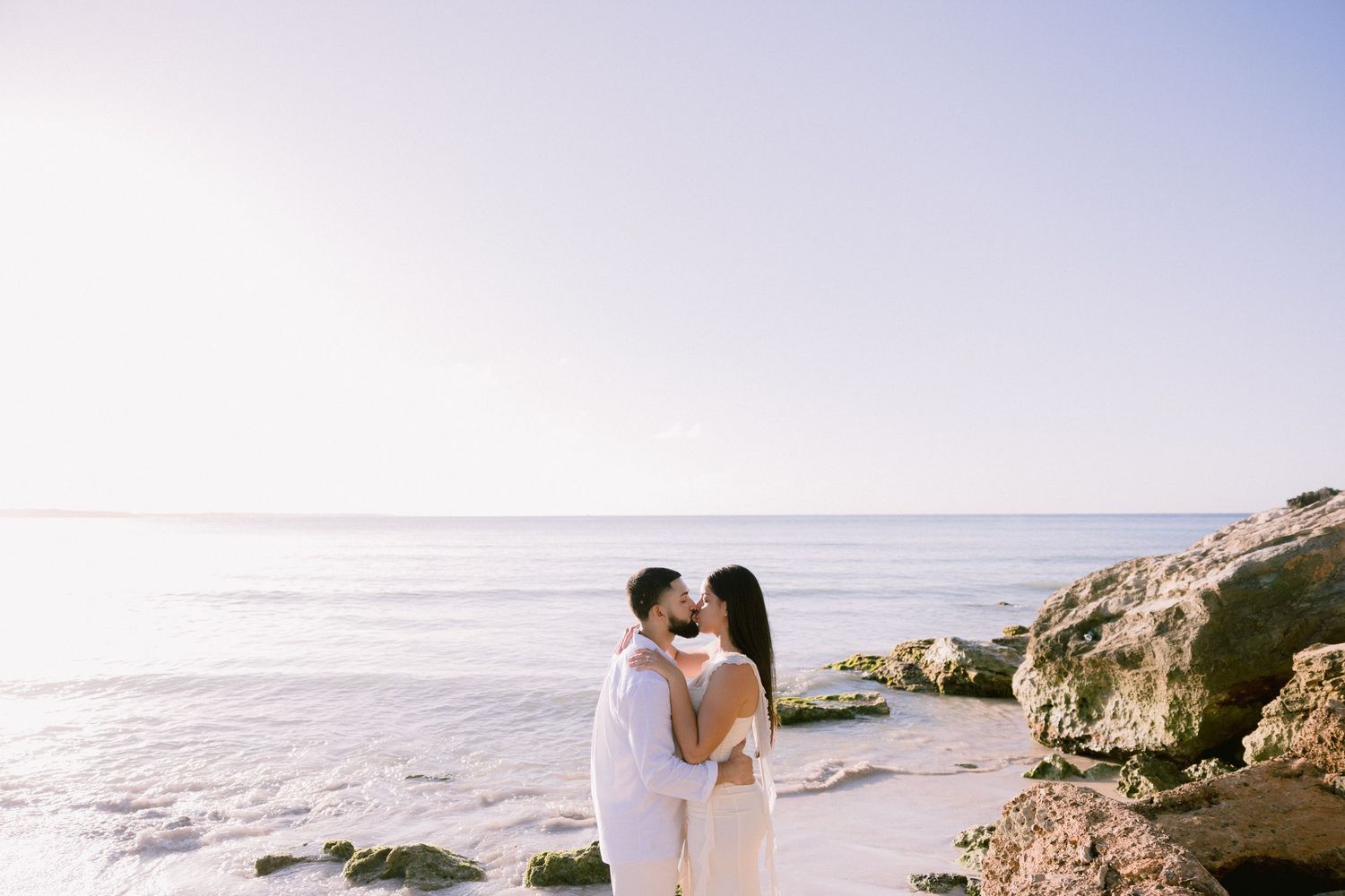 Couple sharing a romantic kiss on a sunny beach with rocky shores and calm ocean waves in the background.