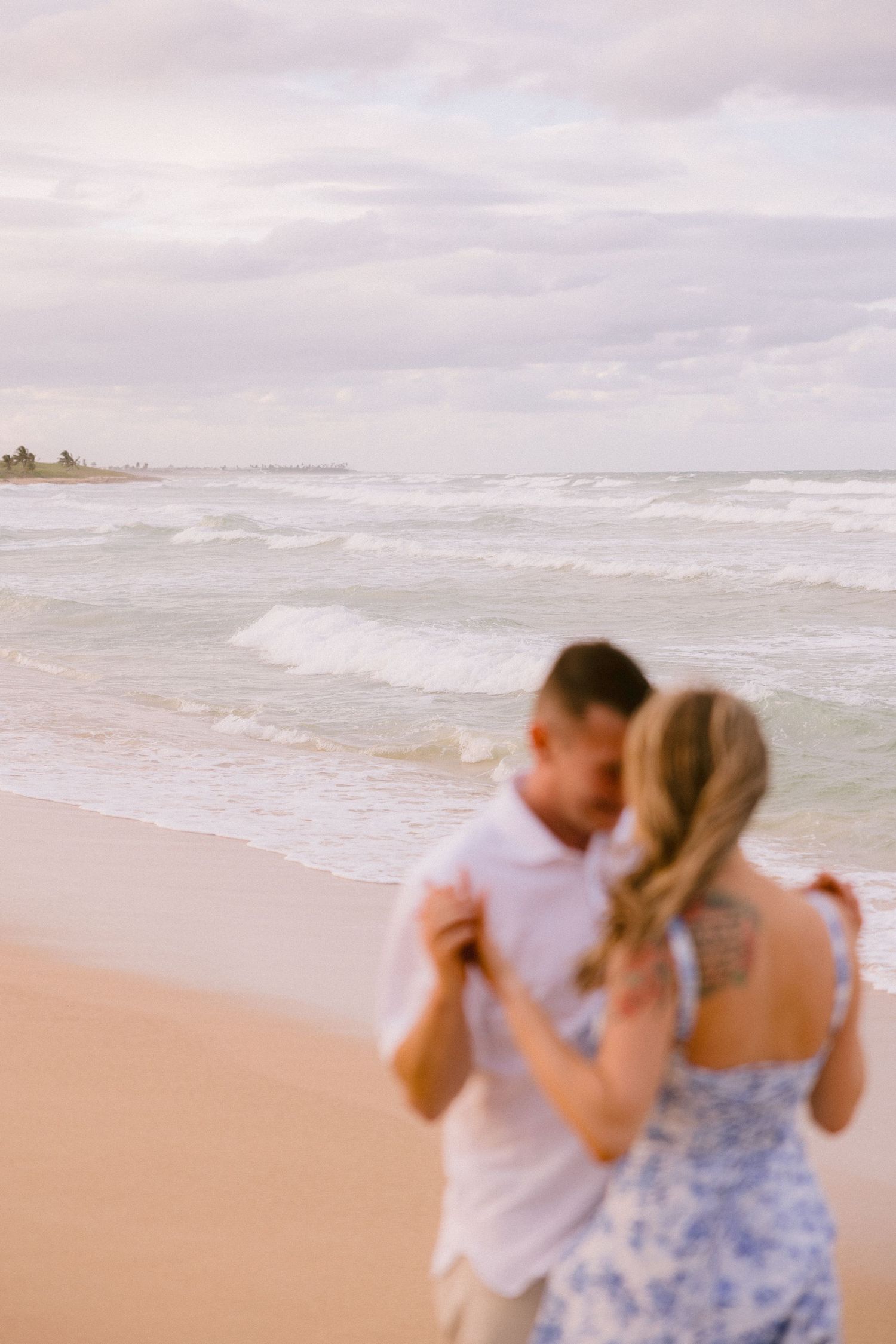 A couple embraces and dances romantically on a sandy beach with ocean waves in the background.