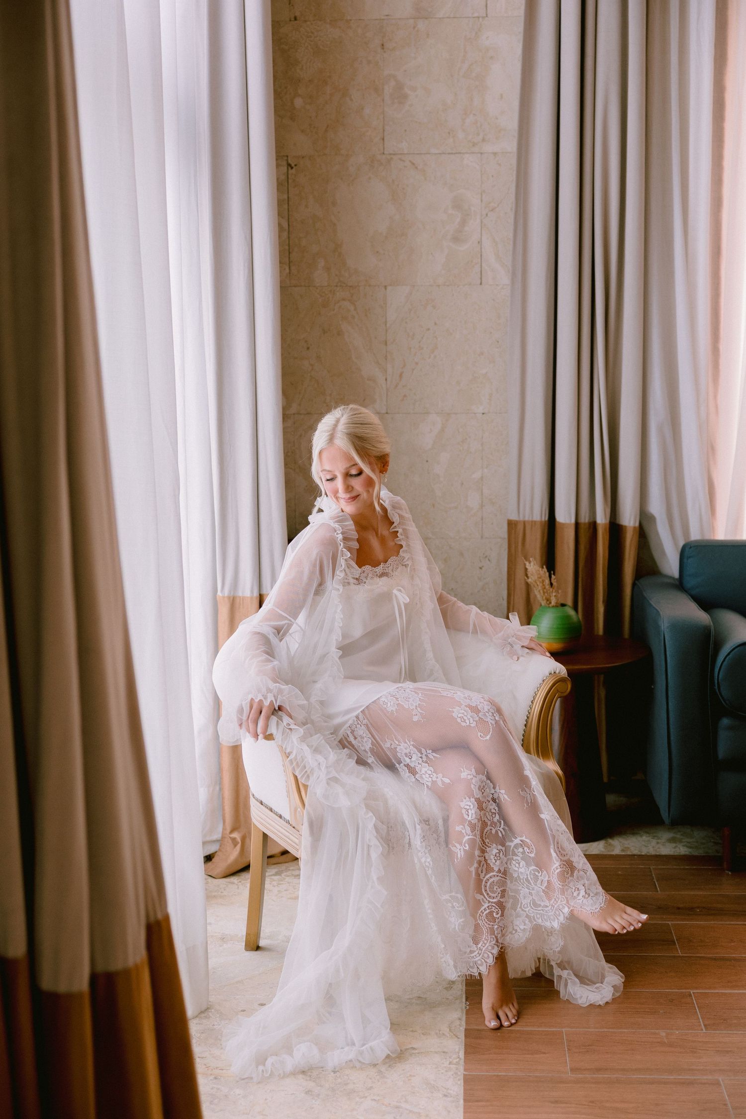 Elegant bride in white lace bridal robe sitting gracefully in a chair by sheer curtains in a luxury hotel room.