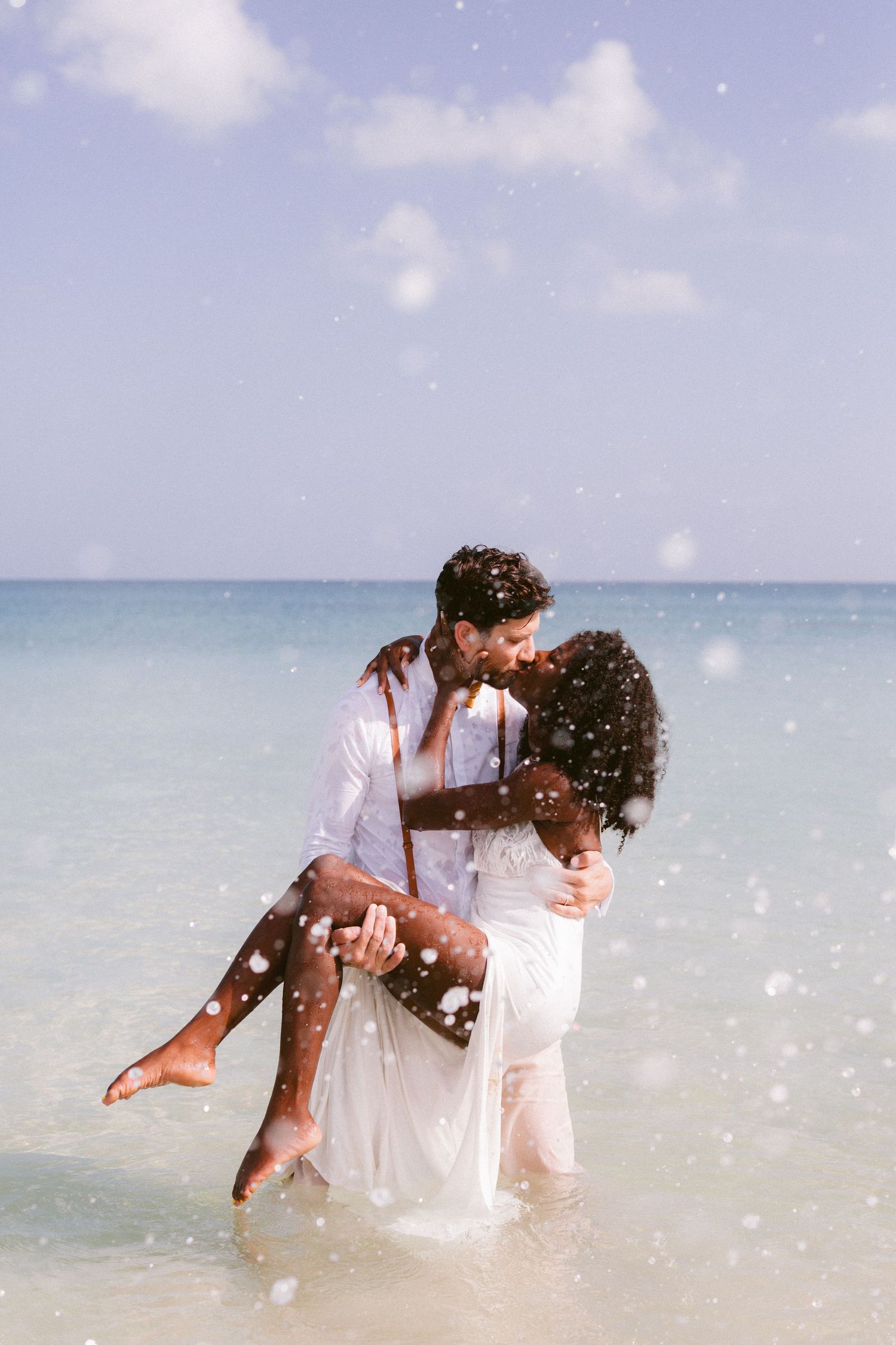 Romantic couple in white outfits kissing on a tropical beach, man carrying woman in shallow ocean water.