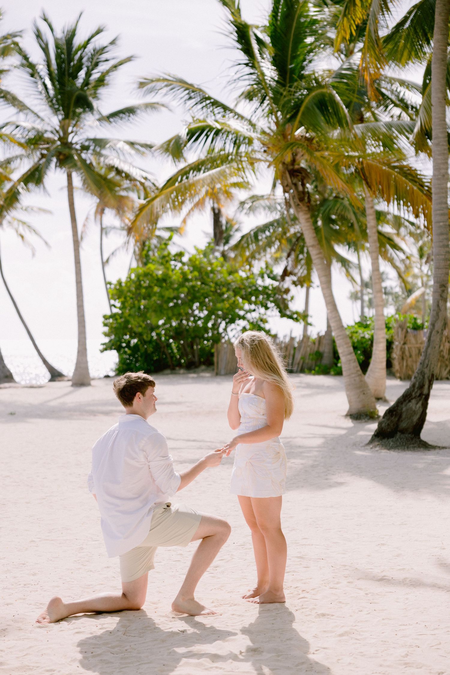 A couple on a tropical beach with palm trees shares a romantic proposal moment on white sand.