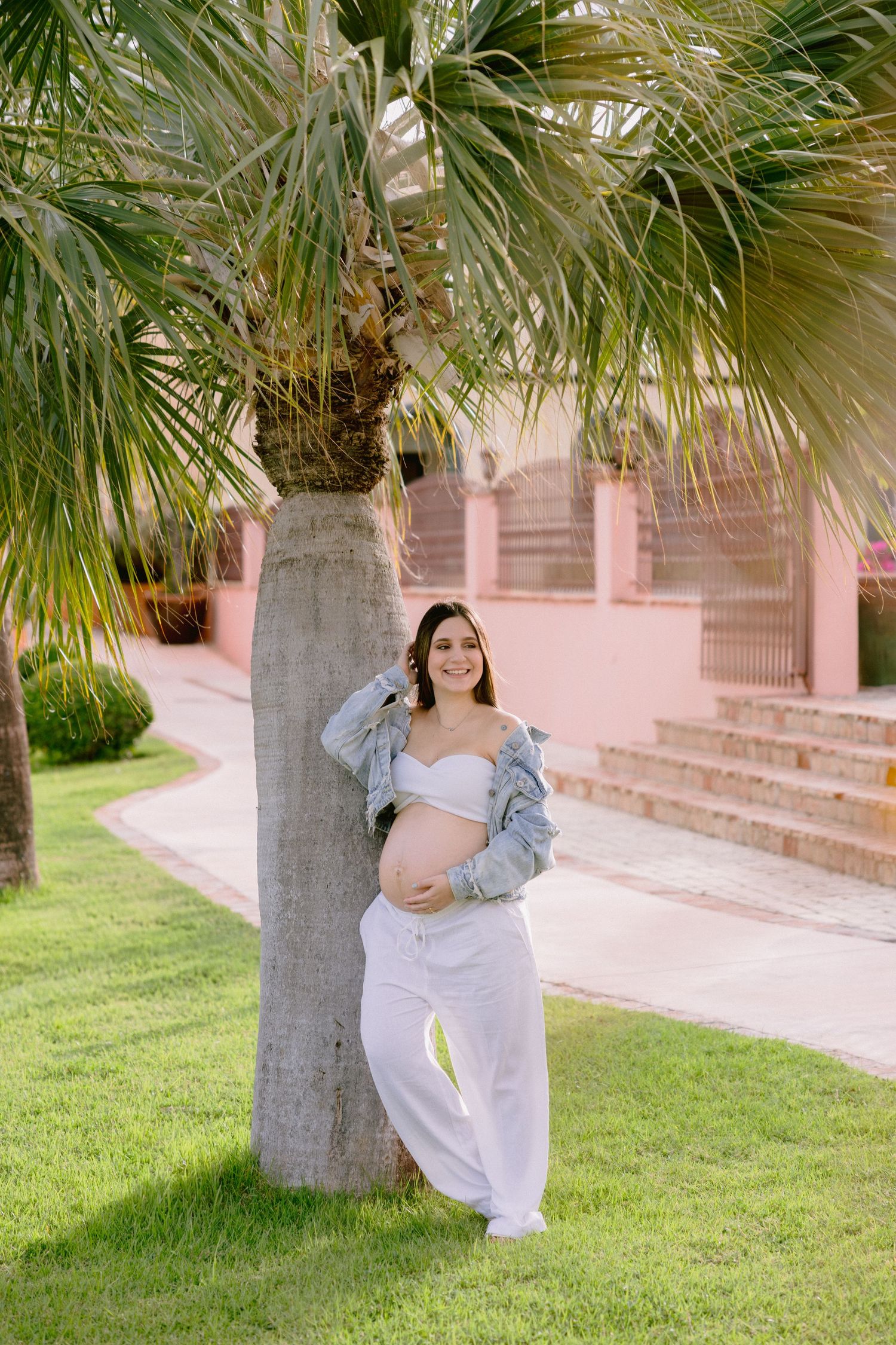 A figure stands near a palm tree wearing casual white pants and a denim jacket on a manicured lawn with pink walls in background.
