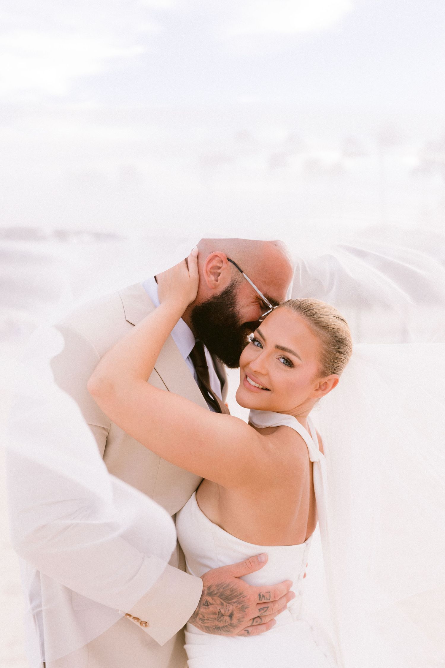 Bride and groom embrace on a beach, with veil flowing, as the smiling bride looks at the camera during their wedding.
