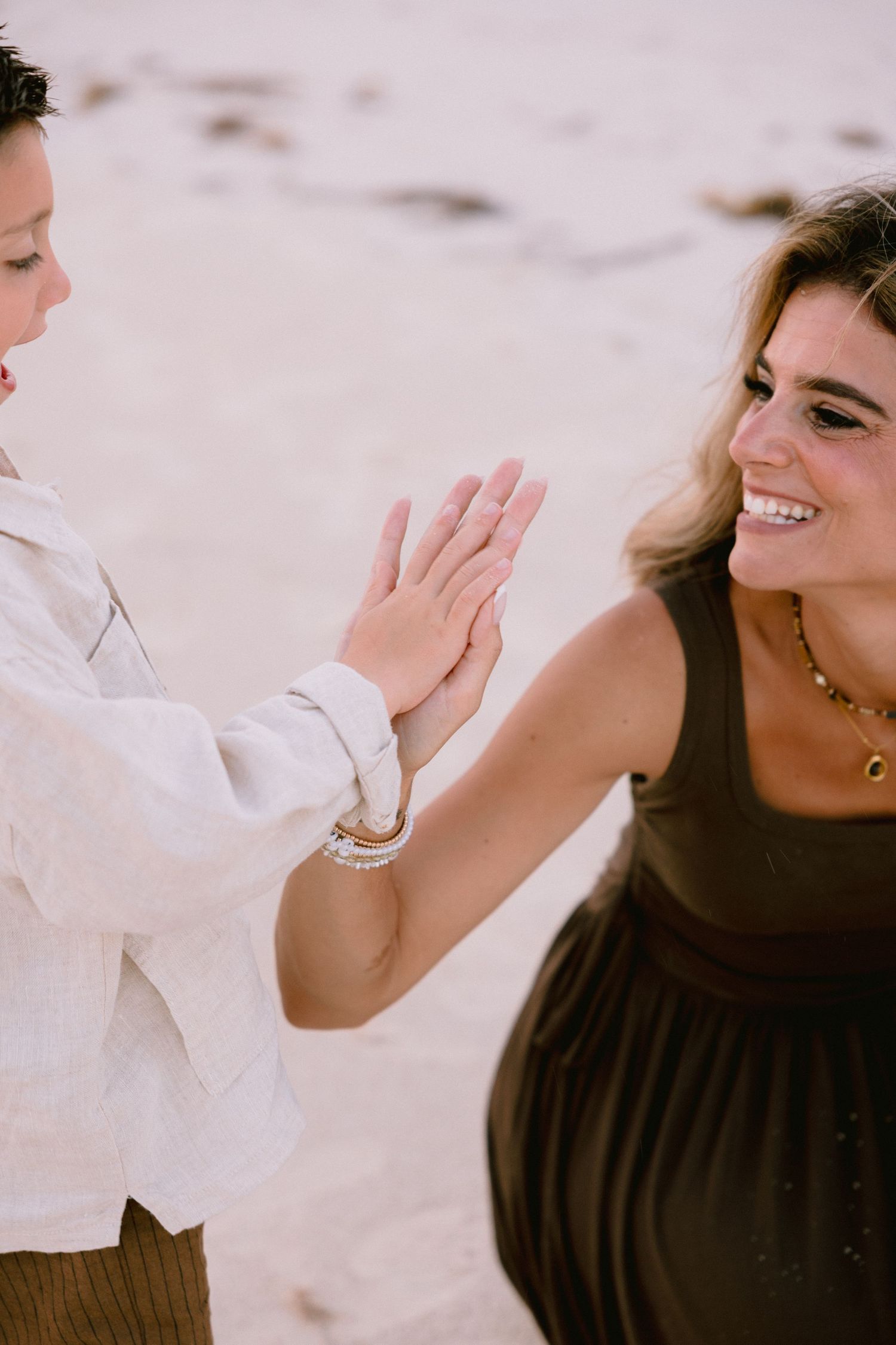 Two women smiling and holding hands on a sandy beach, one wearing a white shirt and the other a dark dress.