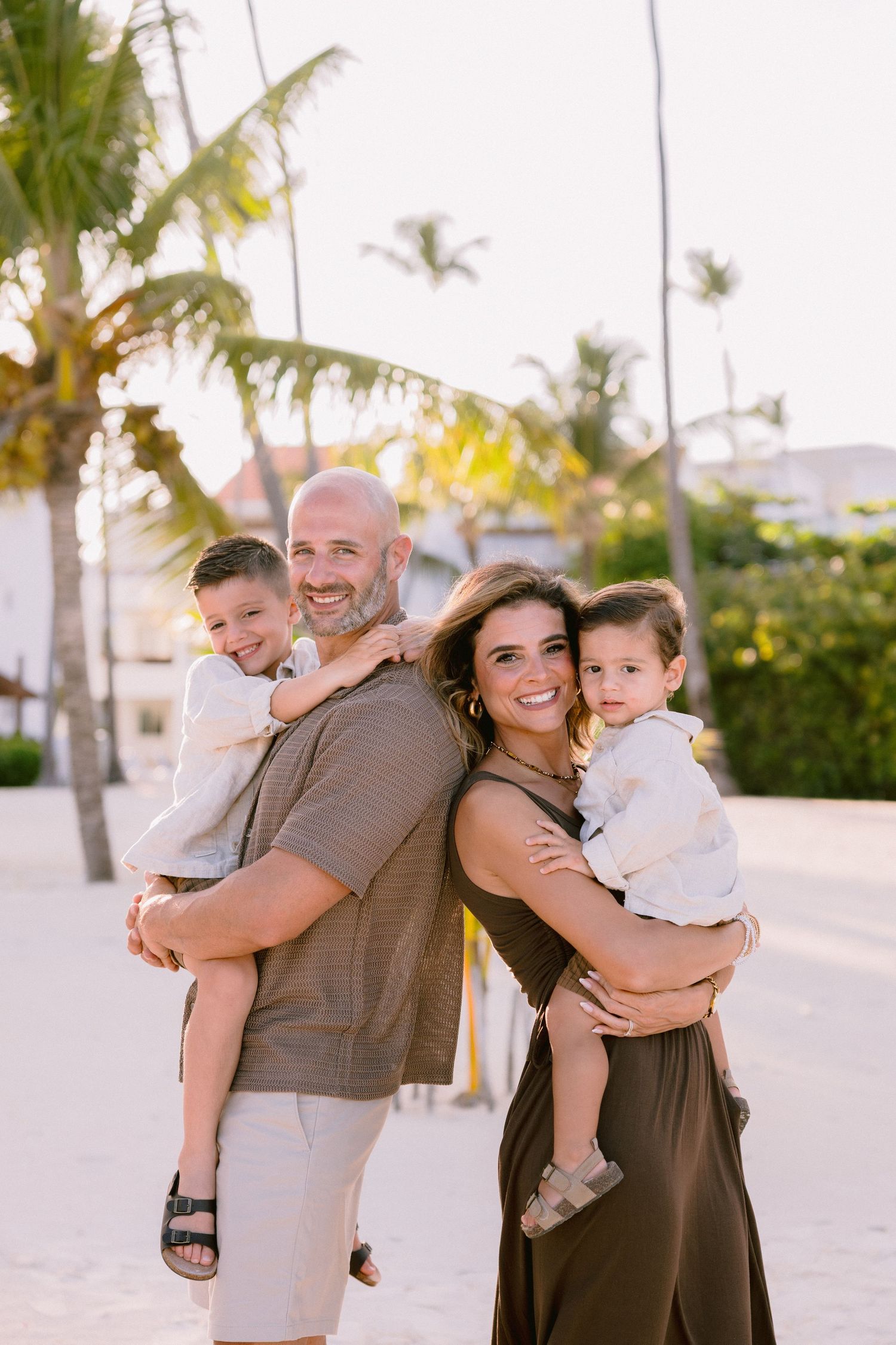 A family embraces and laughs together on a sunny beach with palm trees and white sand in the background.