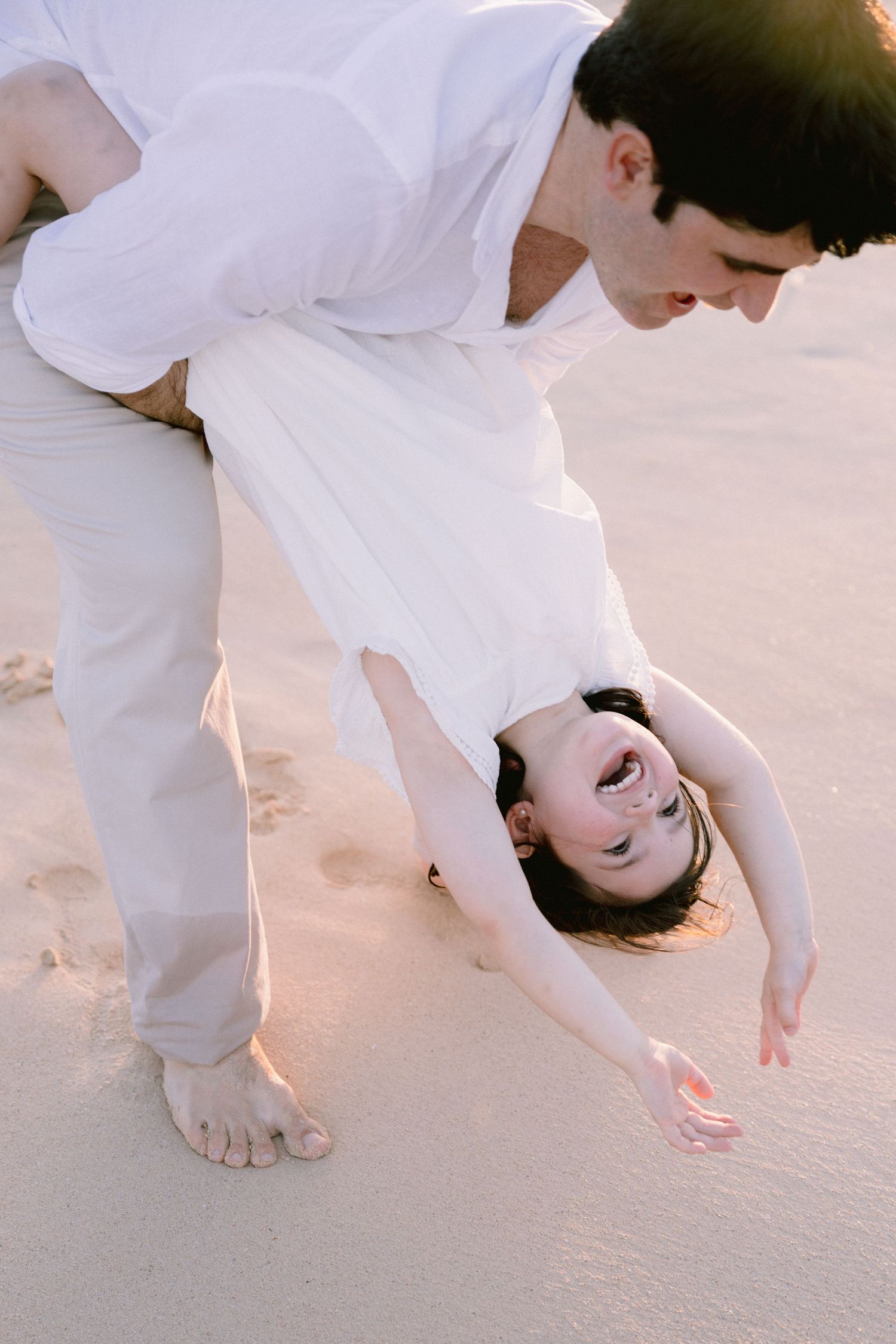 A man in white clothes playfully dips a laughing woman upside down on a sandy beach at sunset.
