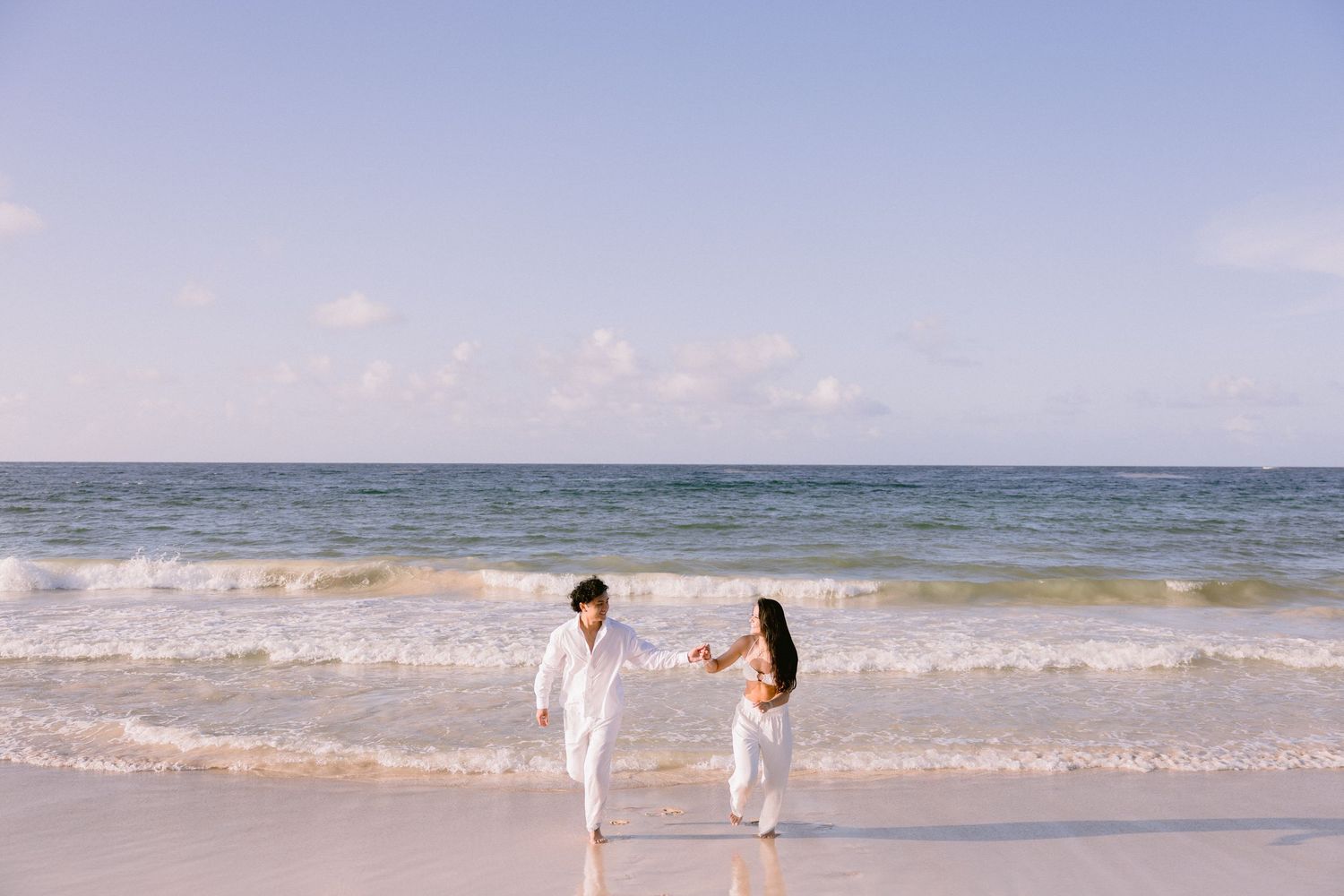 Couple in white outfits holding hands on a sunny beach with ocean waves, romantic beach photoshoot.