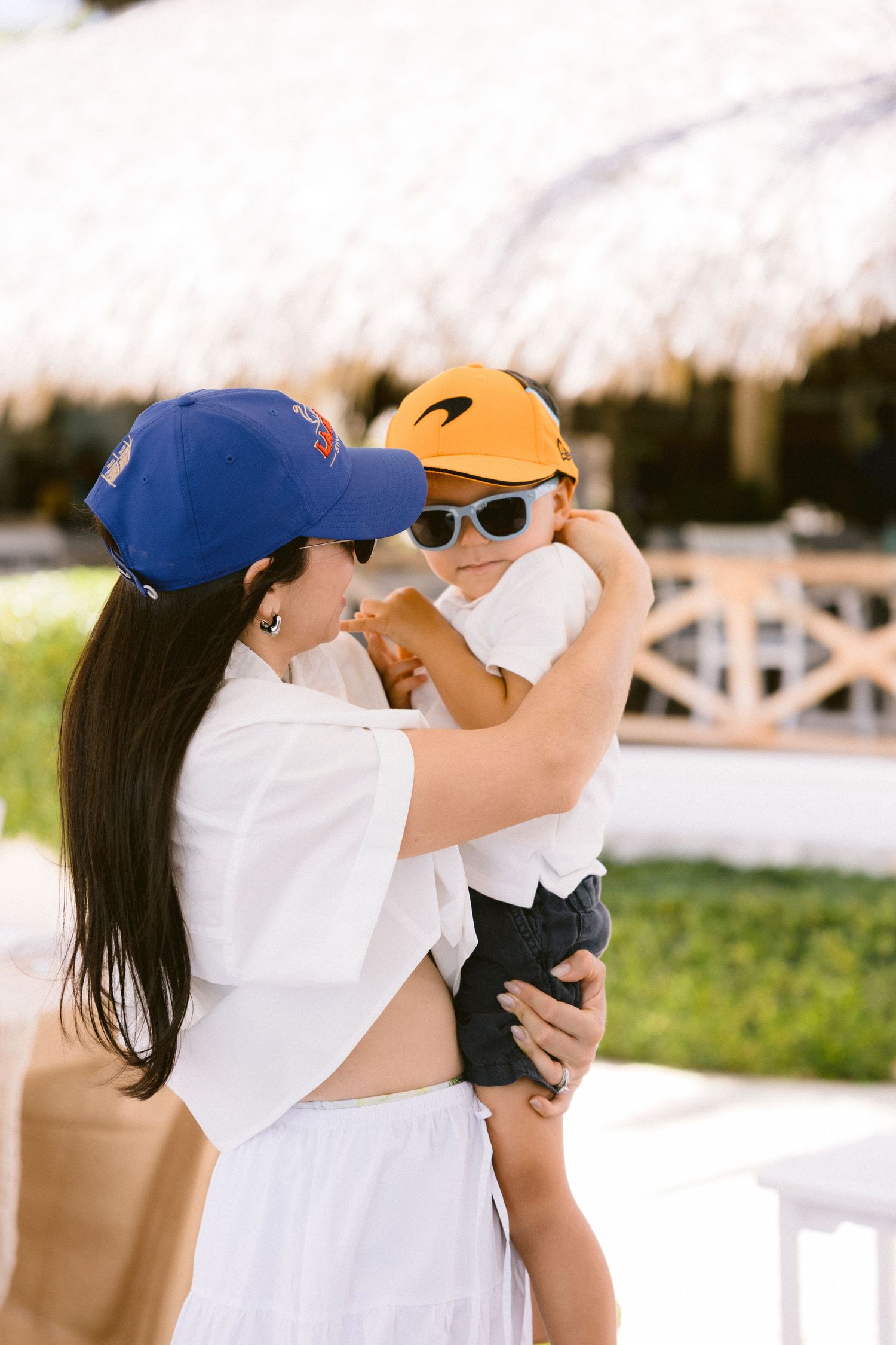 Woman in a blue baseball cap and white outfit holds a smiling toddler in sunglasses and an orange cap near a thatched-roof beach hut.
