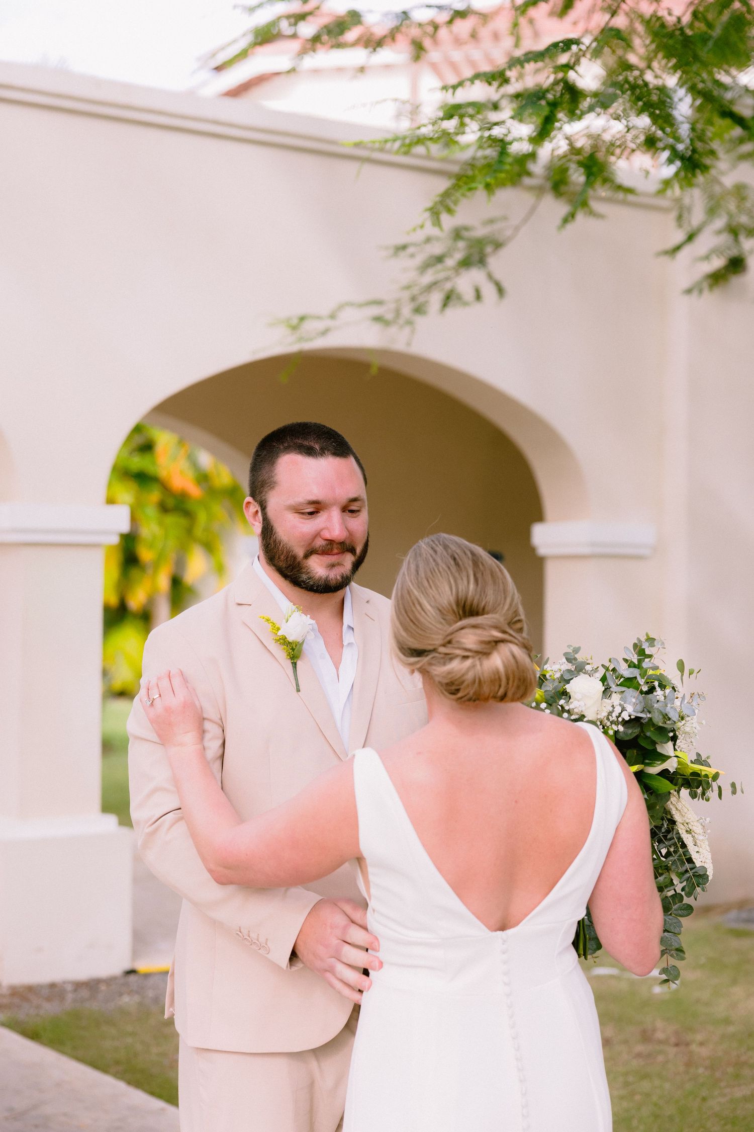 Groom in beige suit smiling at bride in white backless gown holding greenery bouquet outdoors.